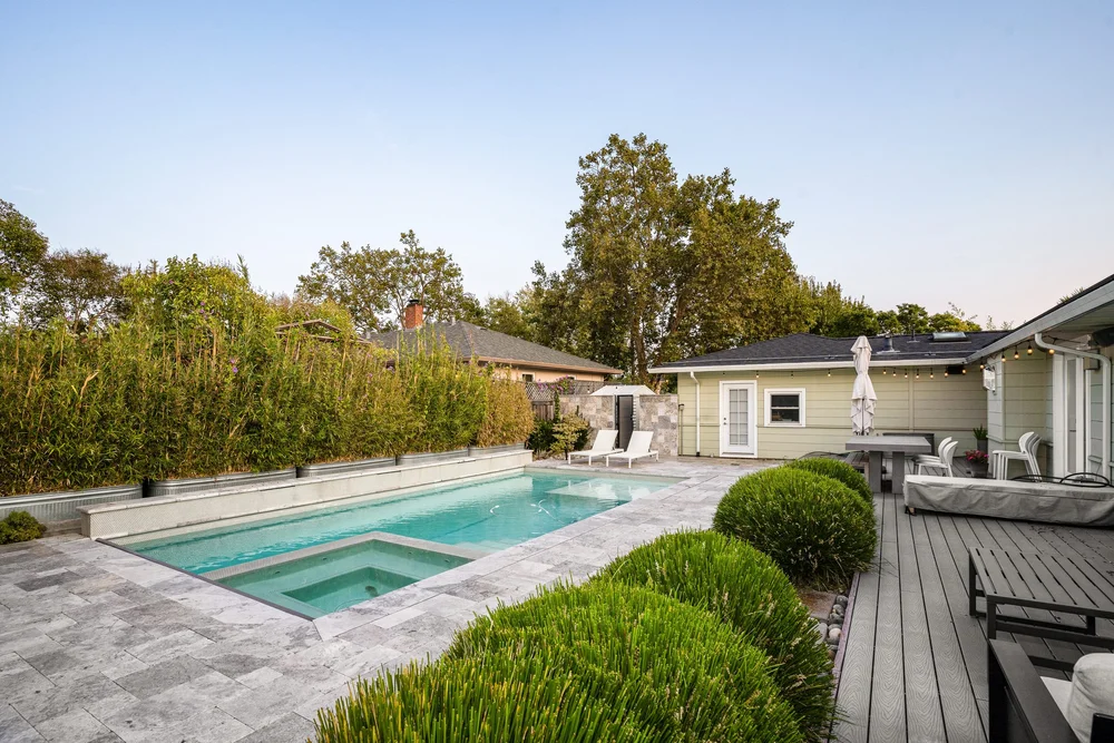Backyard with swimming pool, surrounded by green bushes and trees, patio area with outdoor furniture, and a house in the background.