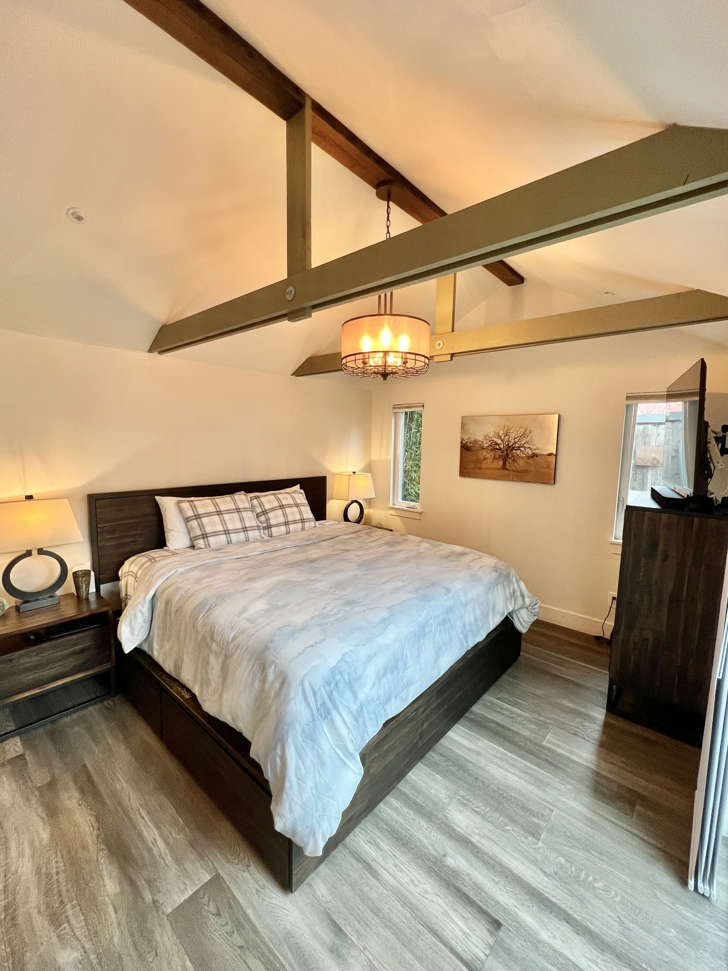 A bedroom with a large bed, bedside lamps, a window on each side of the bed, a ceiling light, and a framed picture of a tree on the wall.