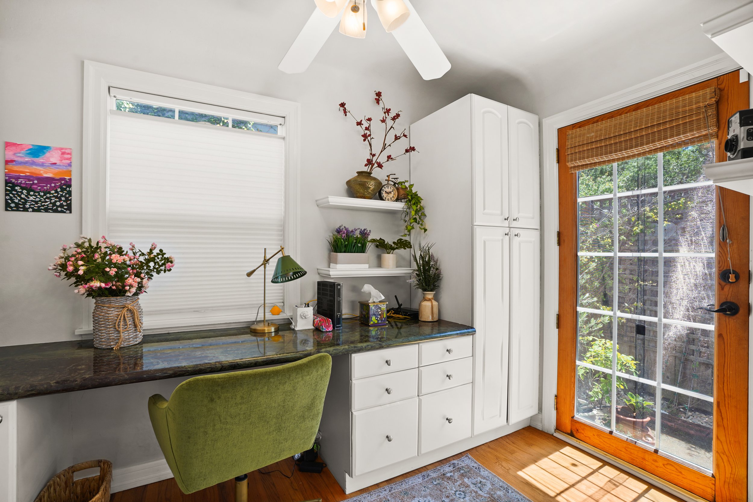 A kitchen counter with a green armchair in front. On the counter, there is a wicker basket with pink flowers, a brass desk lamp, a tissue box, and small electronic devices. White cabinets and a window with a blind are behind the counter. To the side,
