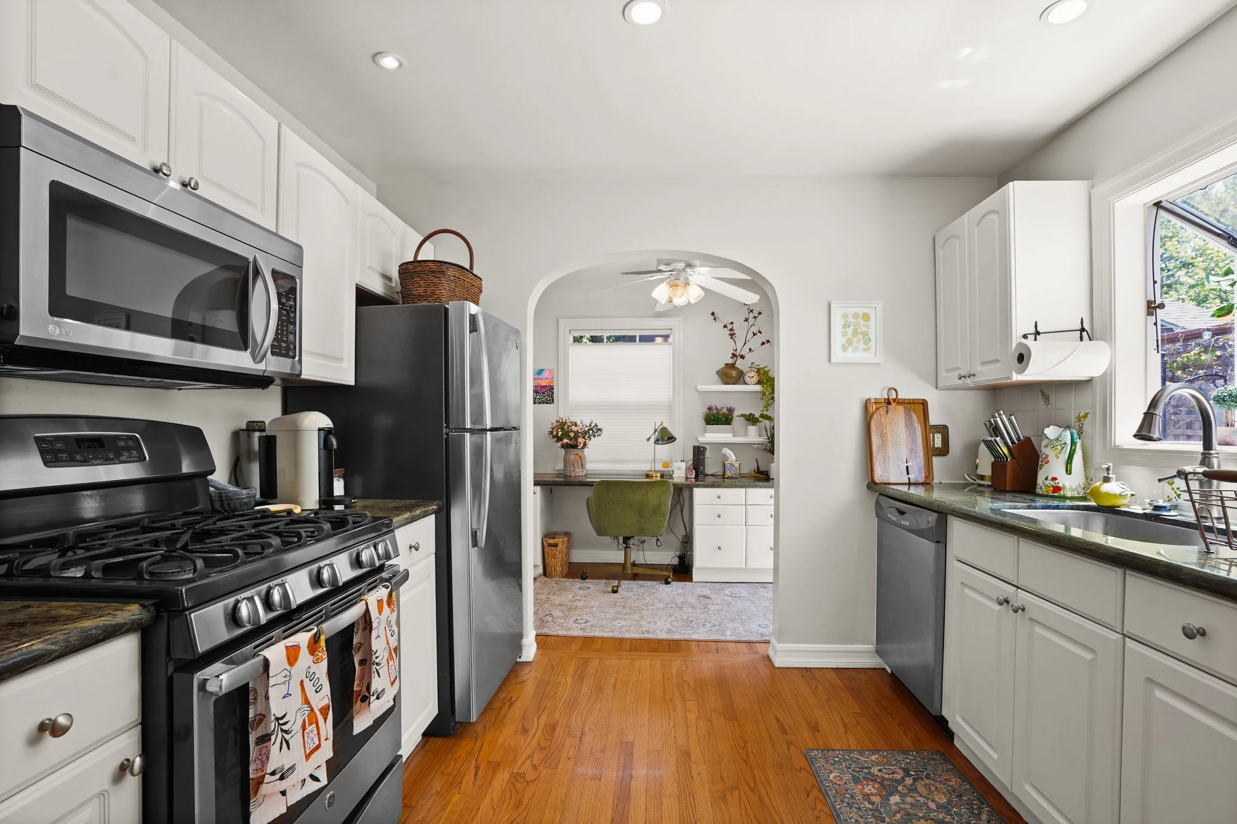 A kitchen with white cabinets, stainless steel appliances including a microwave, refrigerator, and oven, wooden flooring, and a window above the sink showing outdoor greenery. There are countertops with kitchen items, a small rug, and decorative plan