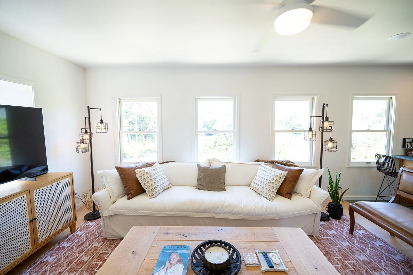 A living room with a white sofa adorned with multiple pillows, situated in front of four windows. Side lamps with cage-style shades, a wooden TV stand with a large flat-screen television, a potted plant, and a brown wooden chair are also visible. A c