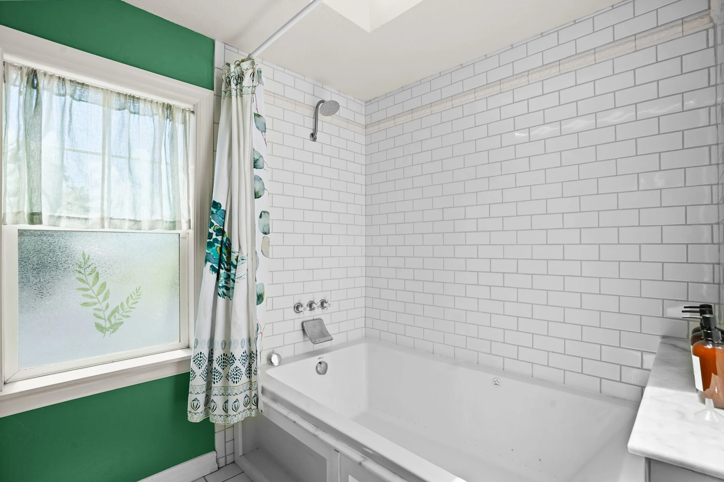 A bathroom with white subway tiles and a bathtub. A window with frosted glass and botanical print curtains is on the left, with green walls around it. Showerhead is mounted on the tiled wall above the tub, and a soap dish is attached to the wall. A b