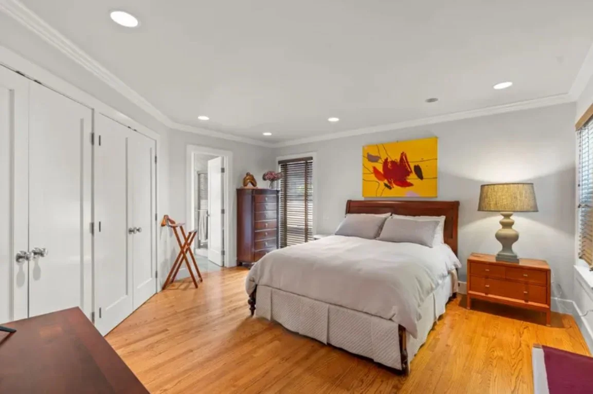 A bedroom featuring a large bed with a wooden headboard, a wooden nightstand with a lamp, a dresser, and closed closets. The room has hardwood flooring, white walls, and is decorated with a colorful abstract painting above the bed.