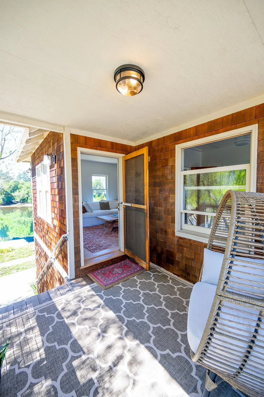 View of a screened porch with a wicker chair, patterned outdoor rug, and a door leading into a sunlit living room with a white sofa and window overlooking greenery.