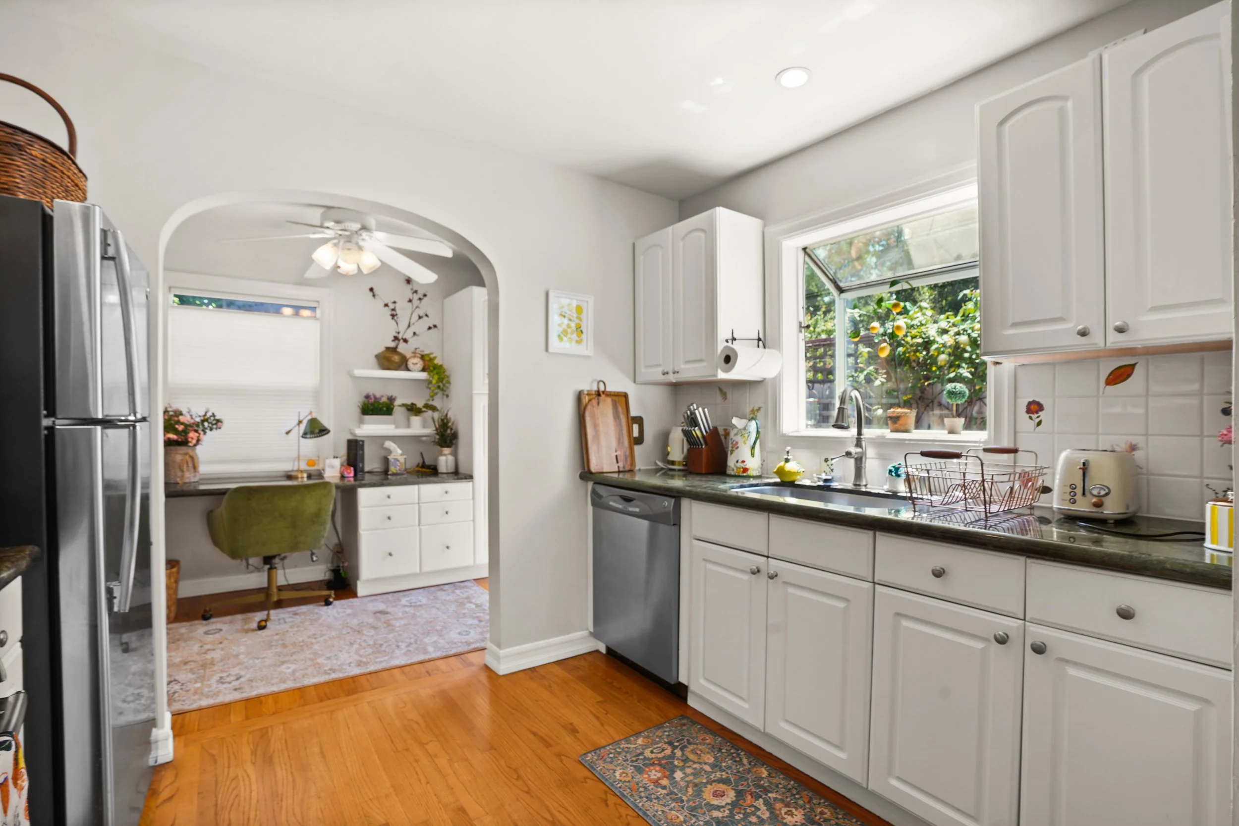 The image shows a cozy kitchen with white cabinets and a large window overlooking a garden. There are various kitchen appliances and items on the black granite countertop, including a dish rack and a toaster. An archway leads to a small office nook w