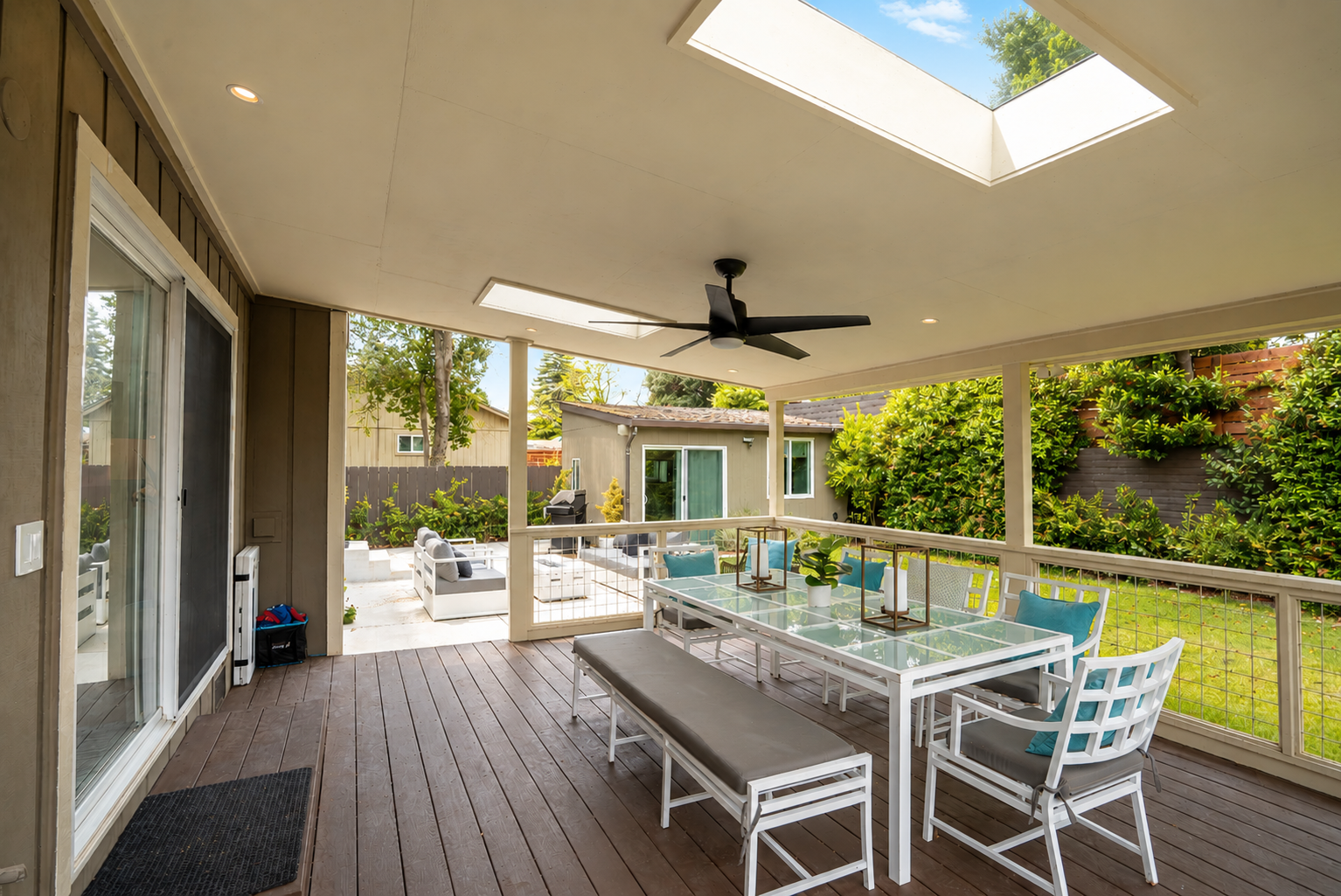 Covered patio with wooden floor, outdoor dining table with chairs, ceiling fan, and skylights, overlooking a backyard with trees, shrubs, and a small building with sliding glass doors.