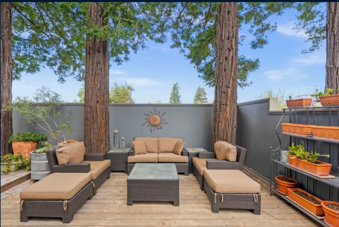 Outdoor patio with gray wicker furniture and beige cushions, surrounded by tall trees and a gray wall, with potted plants and a decorative sun wall hanging.