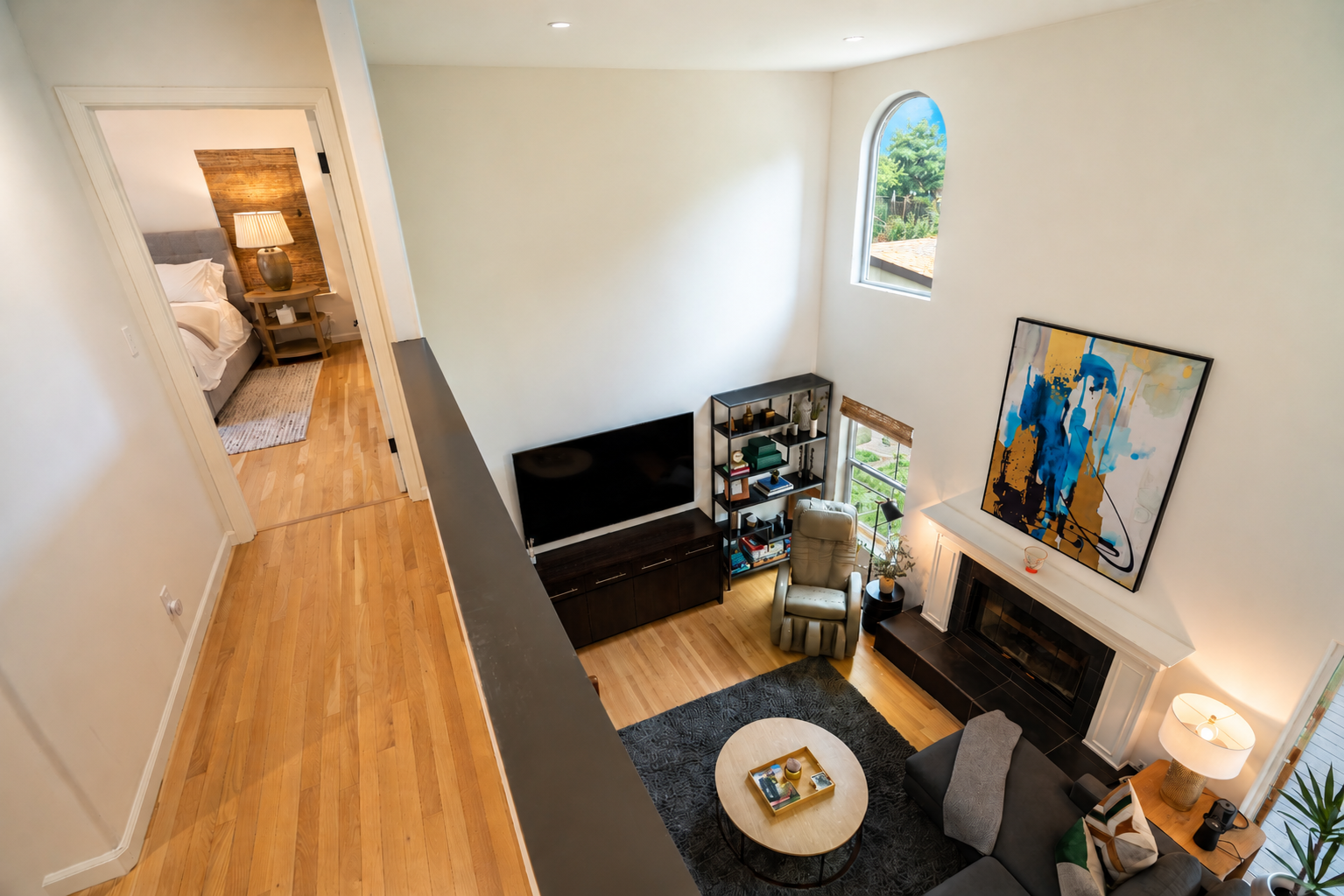 Interior view of a living room with a fireplace, large abstract artwork, a TV, a beige armchair, a black bookshelf, and a TV stand. Part of an adjacent bedroom with a bed and a bedside table with a lamp is visible.