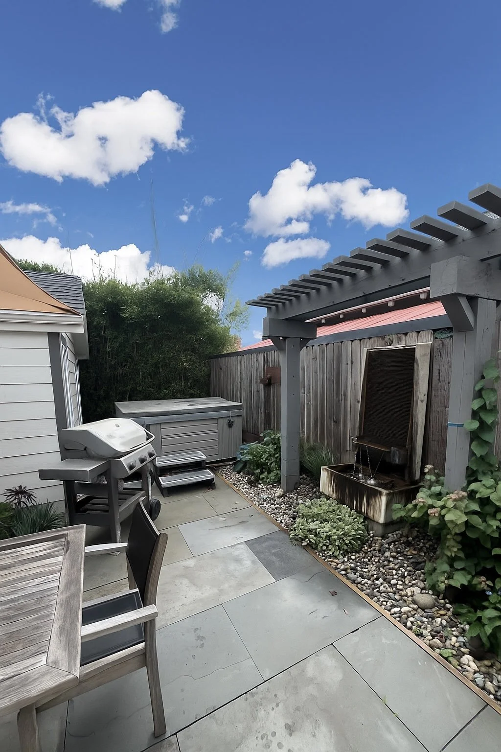 Backyard patio with patio furniture, a barbecue grill, a water feature, and a wooden pergola under a partly cloudy sky.