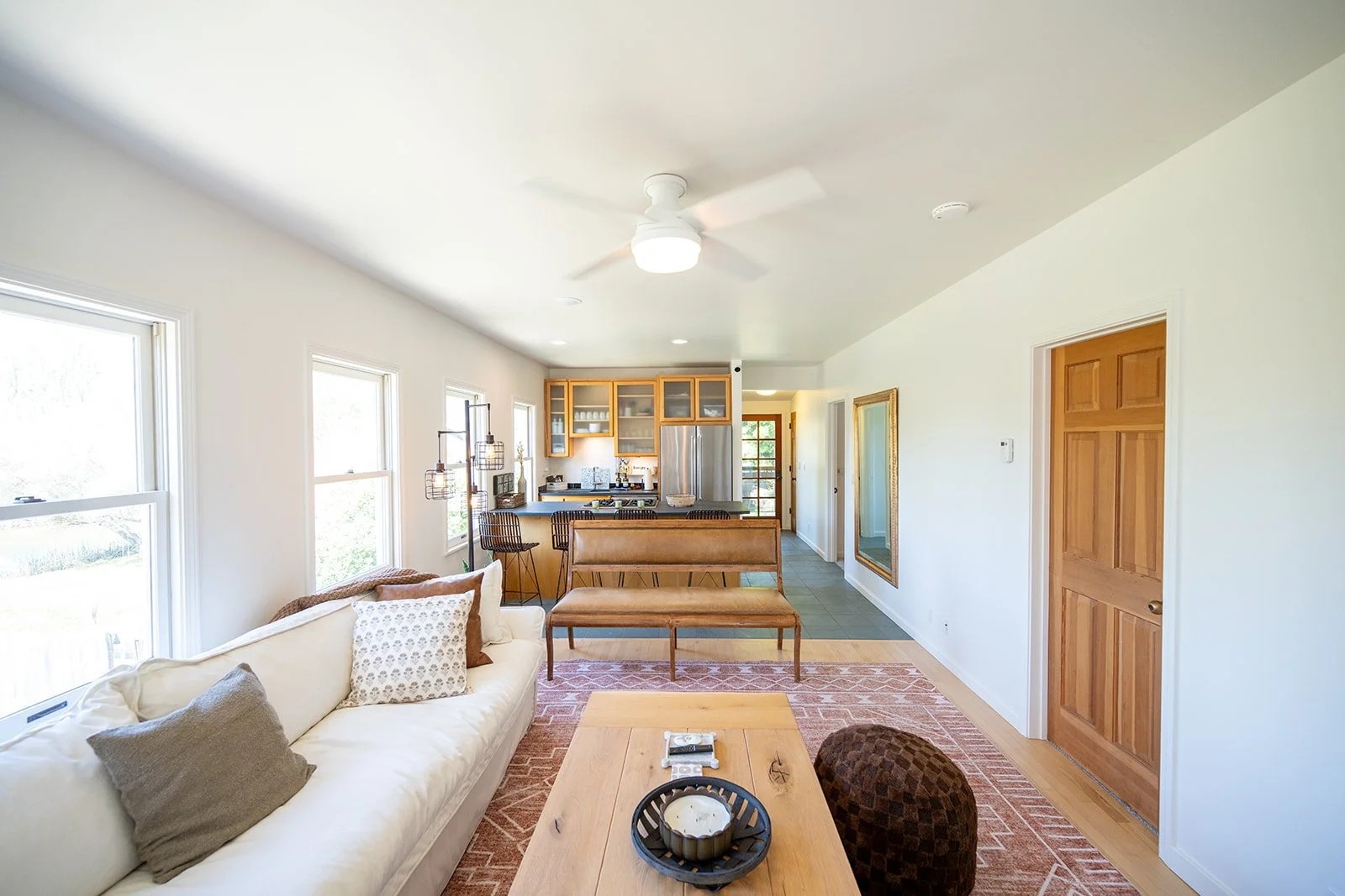 Living room with white sofa, multiple cushions, wooden coffee table, pinkish area rug, and open kitchen with wooden cabinets, stainless steel refrigerator, and bar stools.