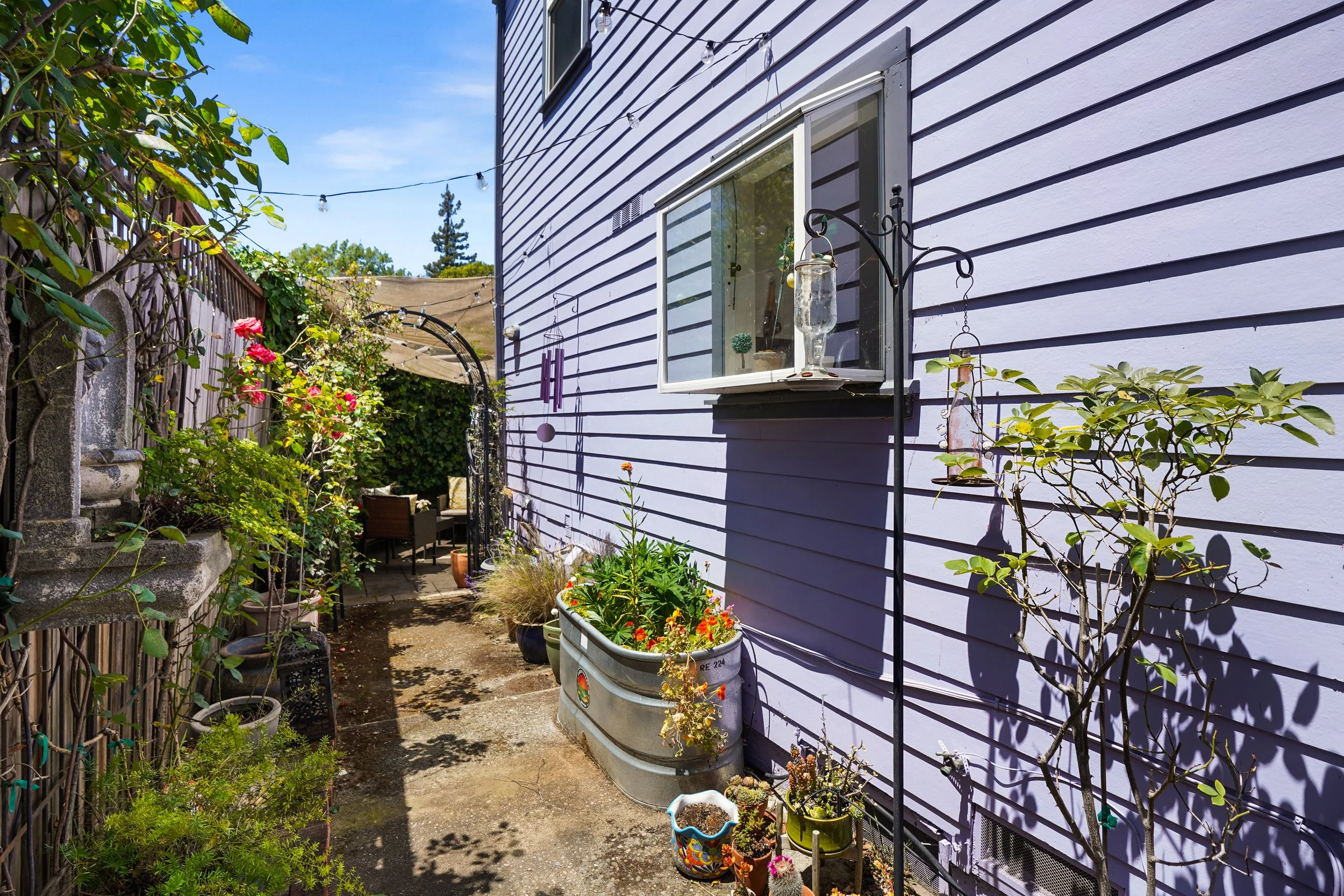 A cozy backyard garden with potted plants, flowers, and outdoor seating, adjacent to a light purple house with horizontal siding.