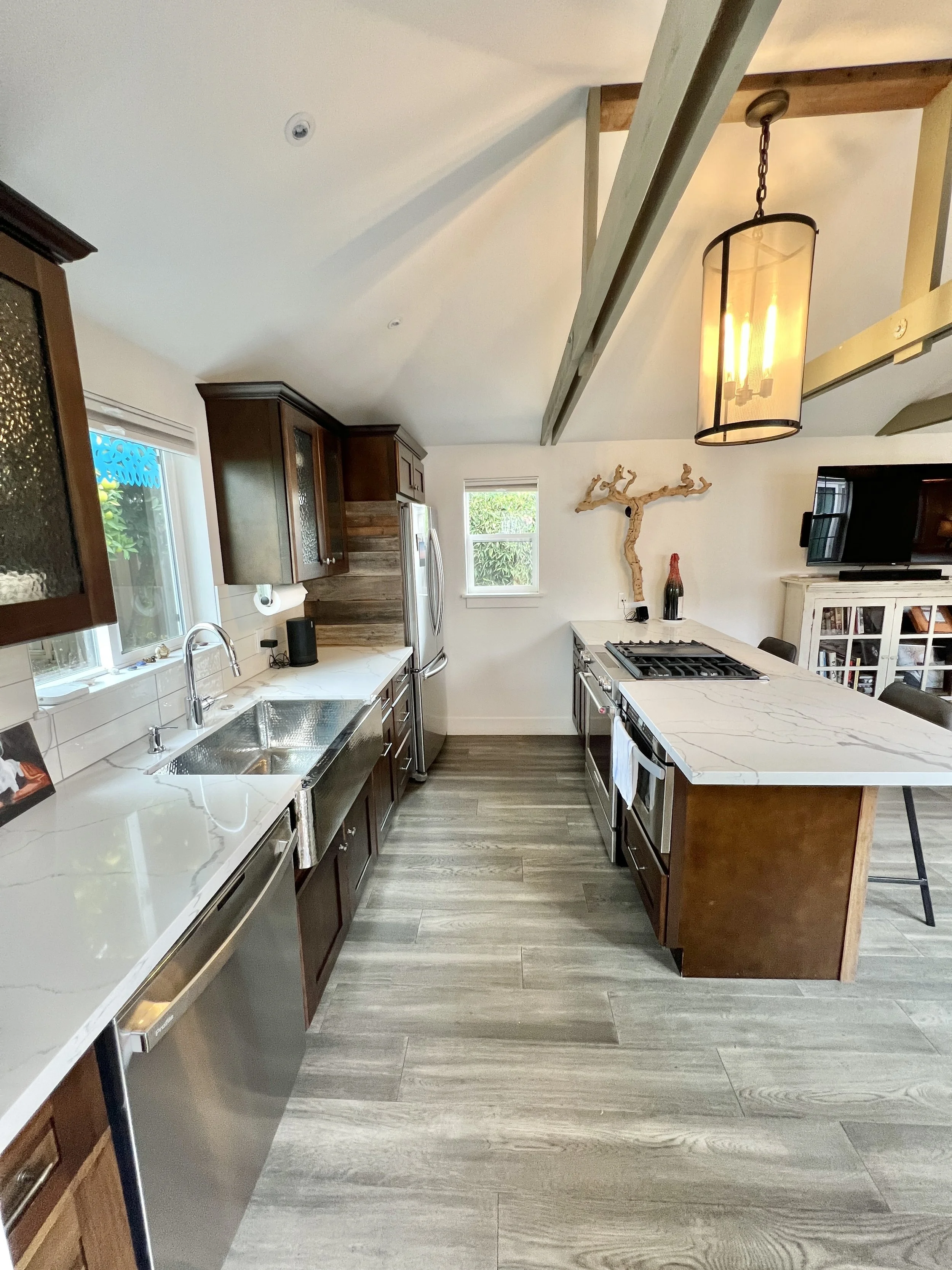 Modern kitchen with white marble countertops, dark wood cabinets, and stainless steel appliances. It features a large island with a marble top, a hanging light fixture, and wooden beams on the ceiling. There are windows letting in natural light and a