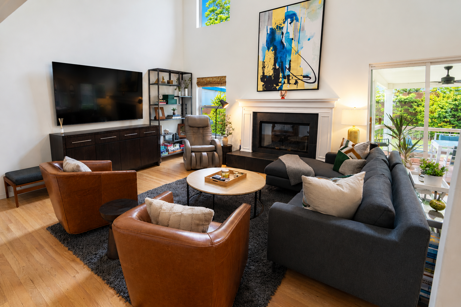 Living room with a large gray sectional sofa, two brown leather armchairs, a round coffee table, a black media console with a flat-screen TV, a beige reclining chair, a white fireplace with a large abstract painting above it, and sliding glass doors 