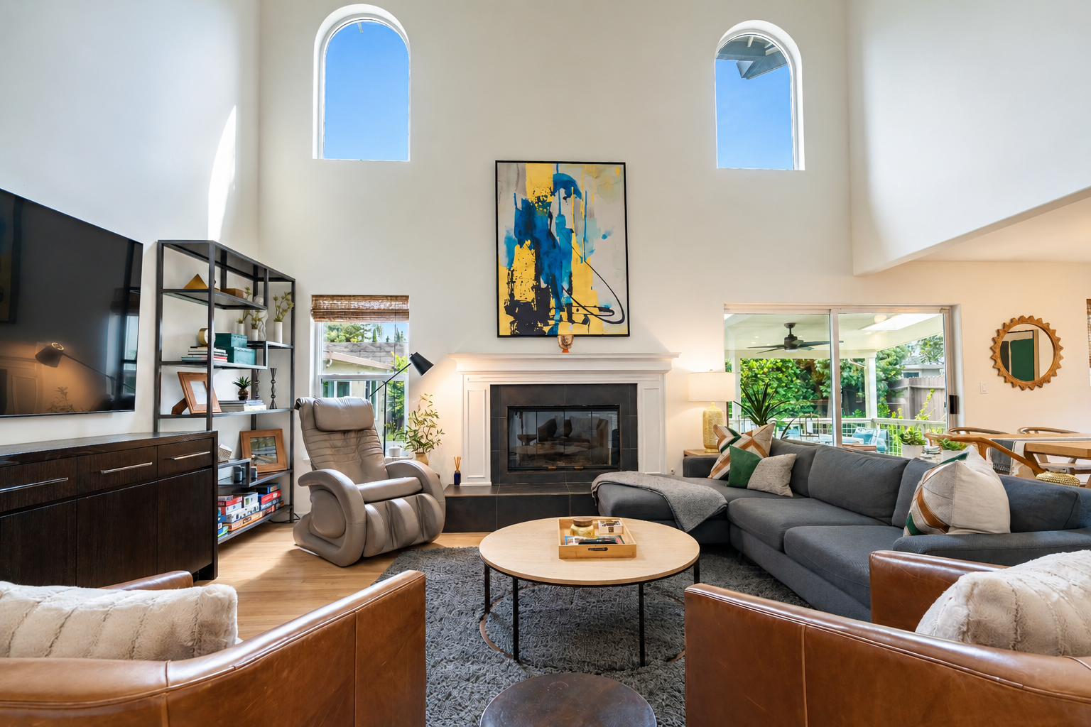Living room with high ceiling, fireplace, gray sectional sofa with pillows, armchair, coffee table, TV, shelves, artwork, large windows, and outdoor view.