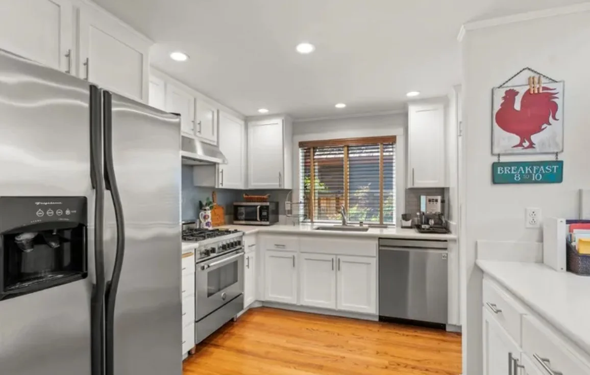 Bright white kitchen with stainless steel appliances, including a refrigerator, stove, microwave, and dishwasher. There's a window with wooden blinds above the sink, hardwood floors, and a wall hanging with a rooster and breakfast hours.