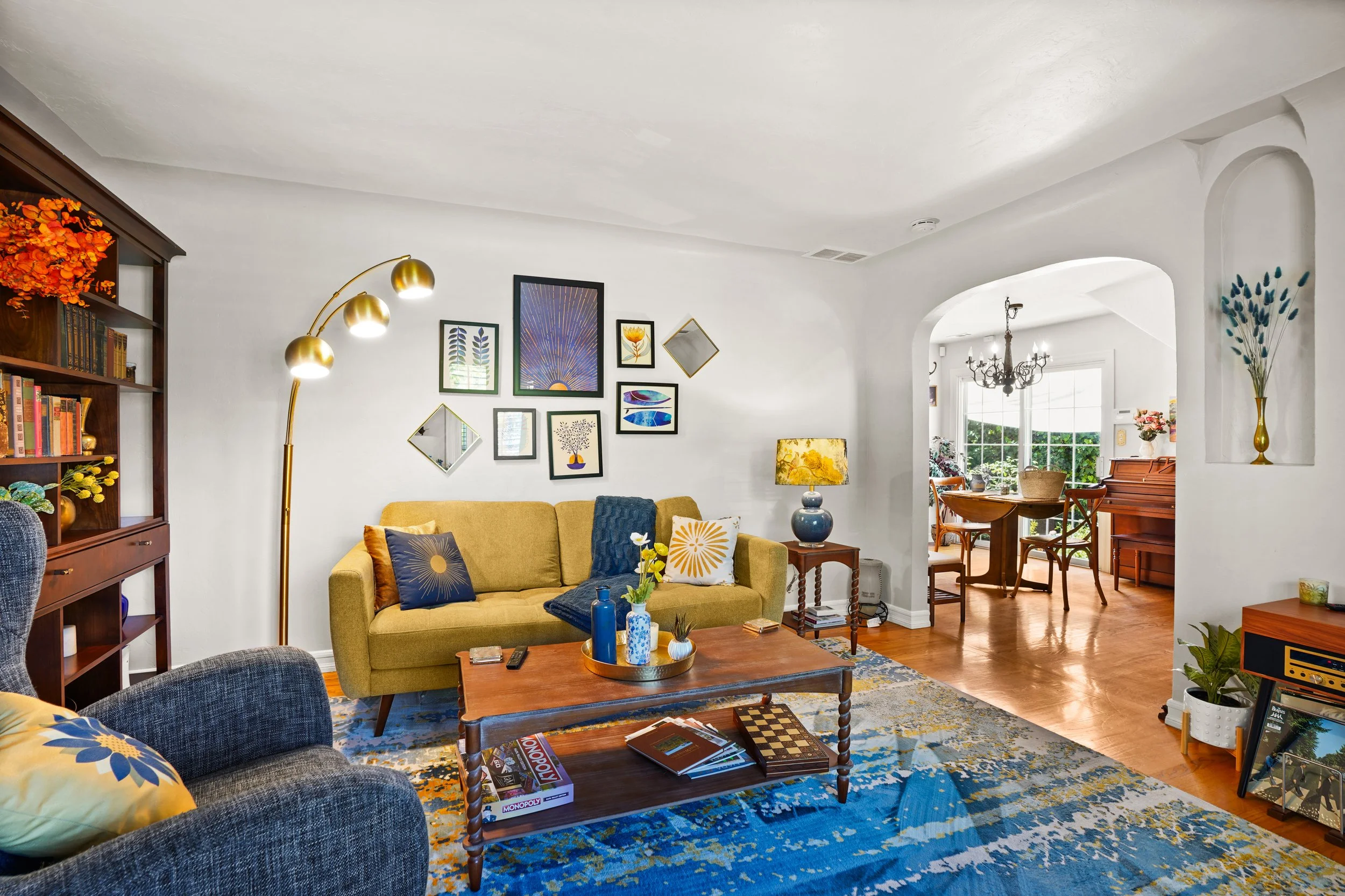 Living room with yellow sofa, dark blue armchair, wooden coffee table, wall art, floor lamp with three gold bulbs, chandelier, and other furniture, with sunlight coming through large windows.