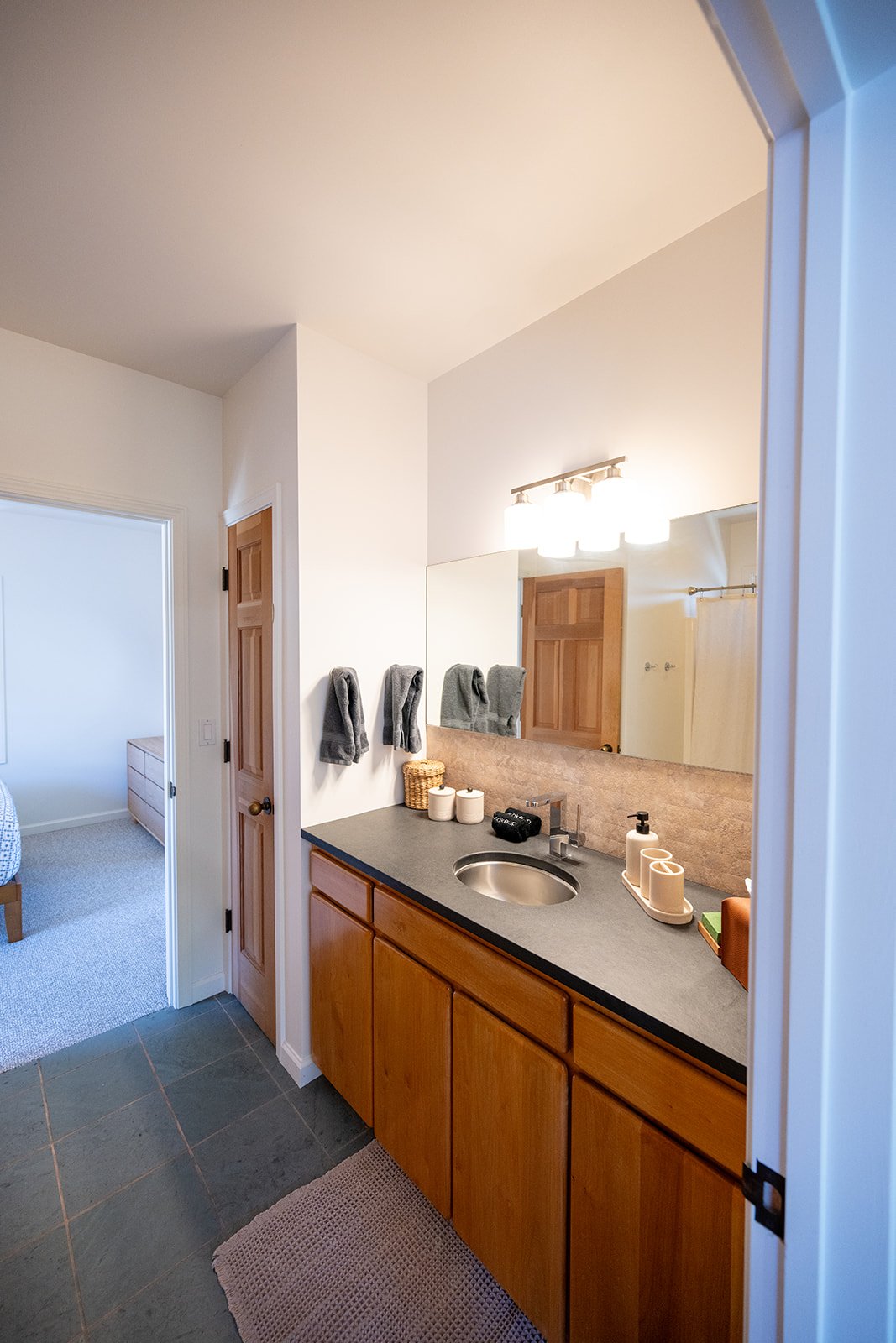 Bathroom vanity with wooden cabinets, gray countertop, stainless steel sink, large mirror, and bathroom accessories such as towels, soap dispenser, and decorative jars.