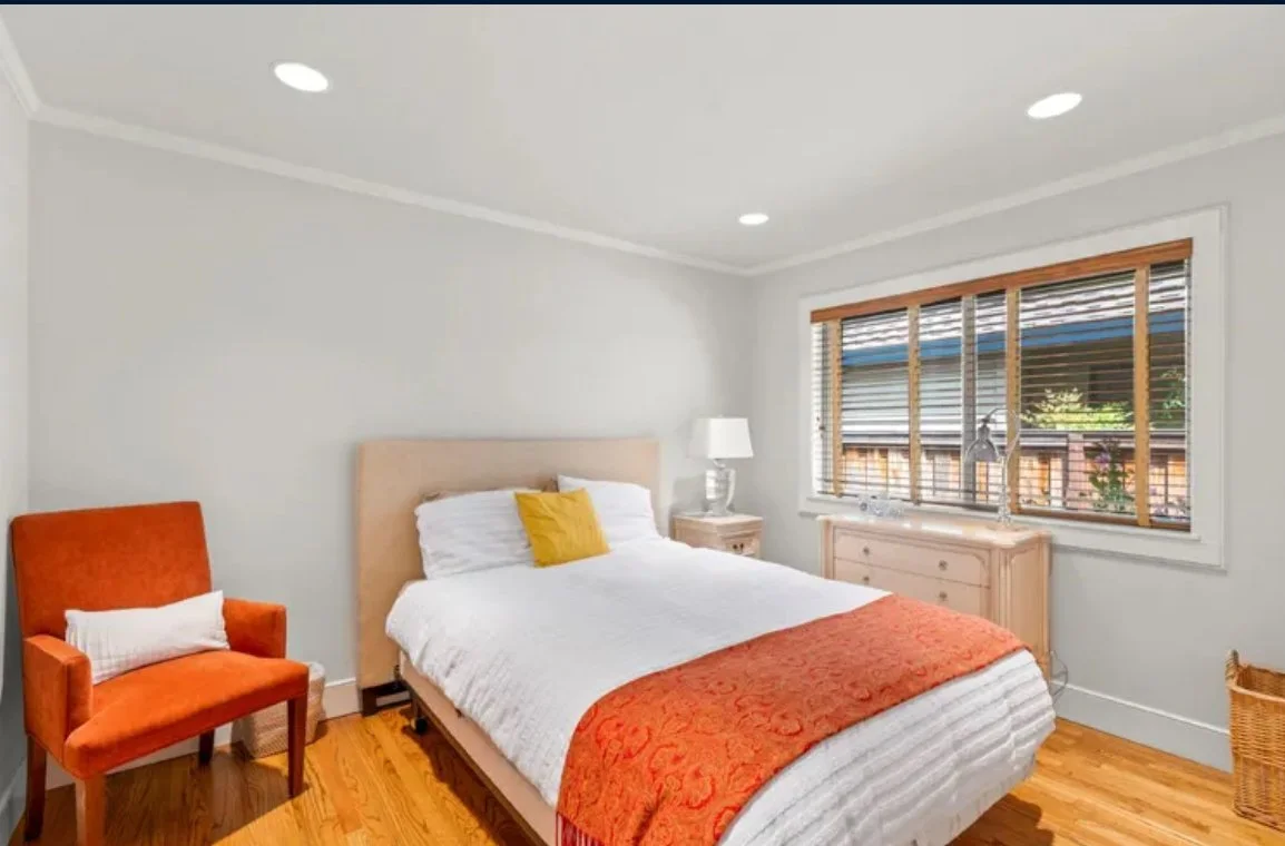 A bedroom with a white bed, orange headboard, yellow throw pillow, orange throw blanket, white nightstand with lamp, orange armchair with pillow, wooden dresser, and large window with wooden blinds