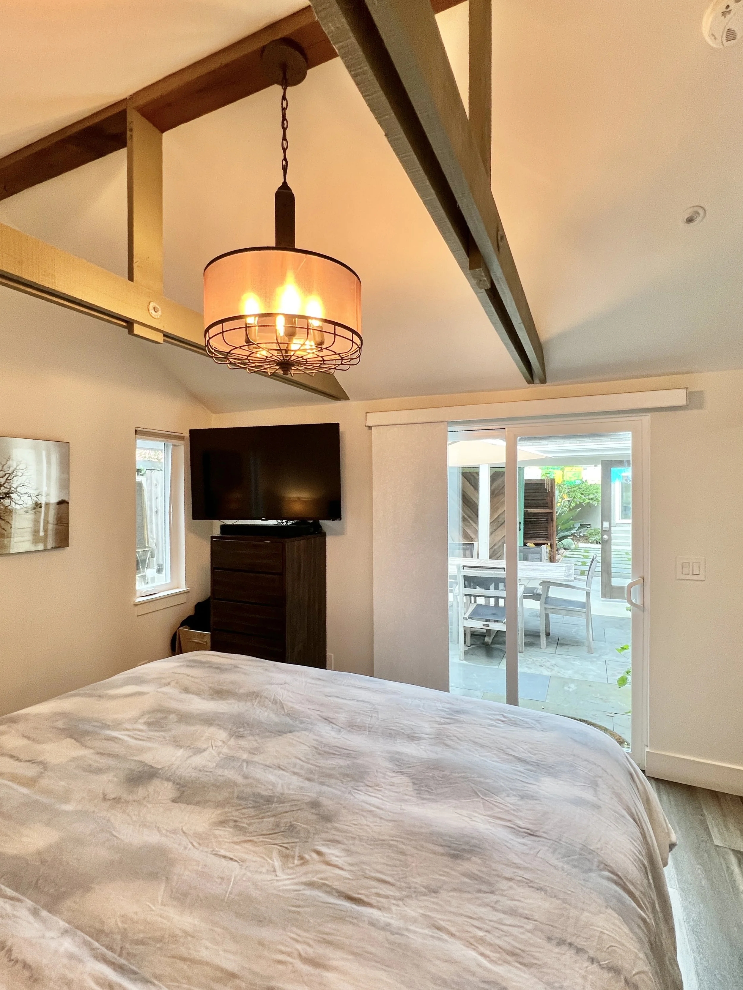A bedroom with a ceiling light fixture, a large bed with a beige bedspread, a wall-mounted TV on a wooden dresser, a small window near the bed, and sliding glass door leading to an outdoor patio with a dining table and chairs.