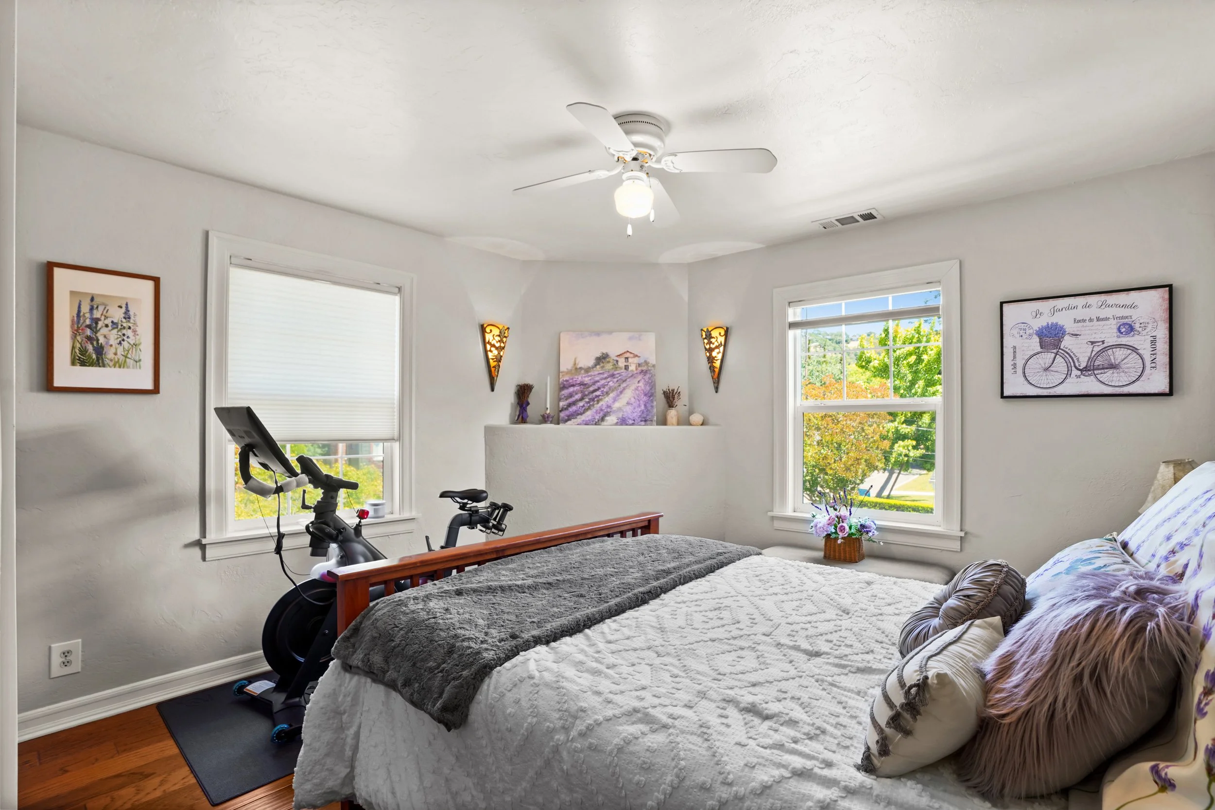 A bedroom with a wooden bed frame, a white quilt, and decorative pillows. There is an exercise bike near the window, which shows a view of trees and a house outside. The room has white walls, two windows, and framed artwork on the walls, with a ceili
