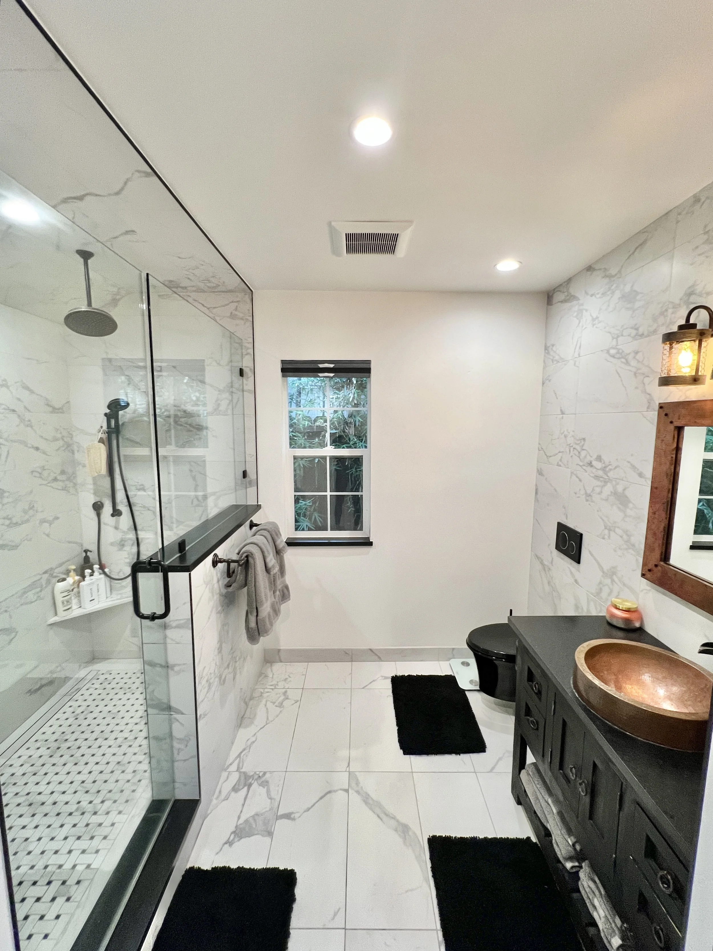 Modern bathroom with a walk-in shower, marble tile walls, a black vanity with a copper sink, black rugs, and a small window.