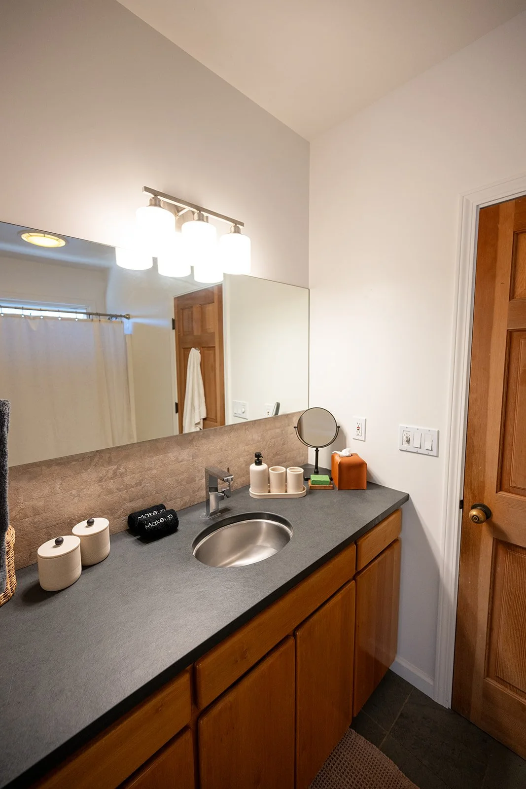 Bathroom vanity with a gray countertop, wooden cabinets, a small round mirror, and various toiletries, with a large mirror and light fixture above.