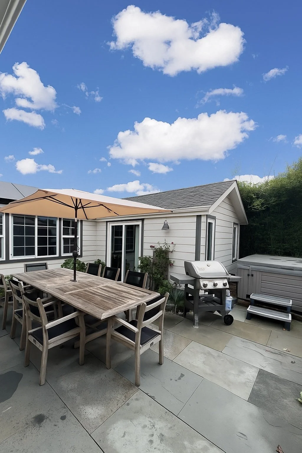 Outdoor patio with wooden dining table, black chairs, large beige umbrella, grill, small hot tub, and a white house with multiple windows and sliding door, under partly cloudy blue sky.
