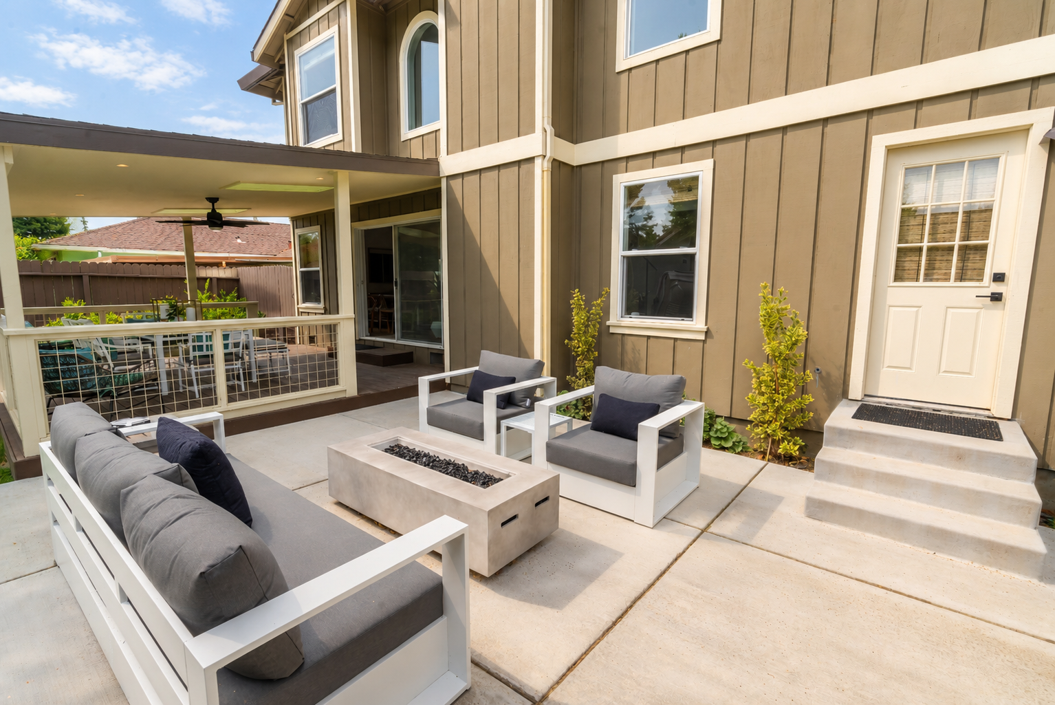 Patio outdoor seating area with cushioned chairs and a fire feature, adjacent to a house with tan siding and white trim, under a covered porch with ceiling fan, overlooking a backyard with a fence and trees.