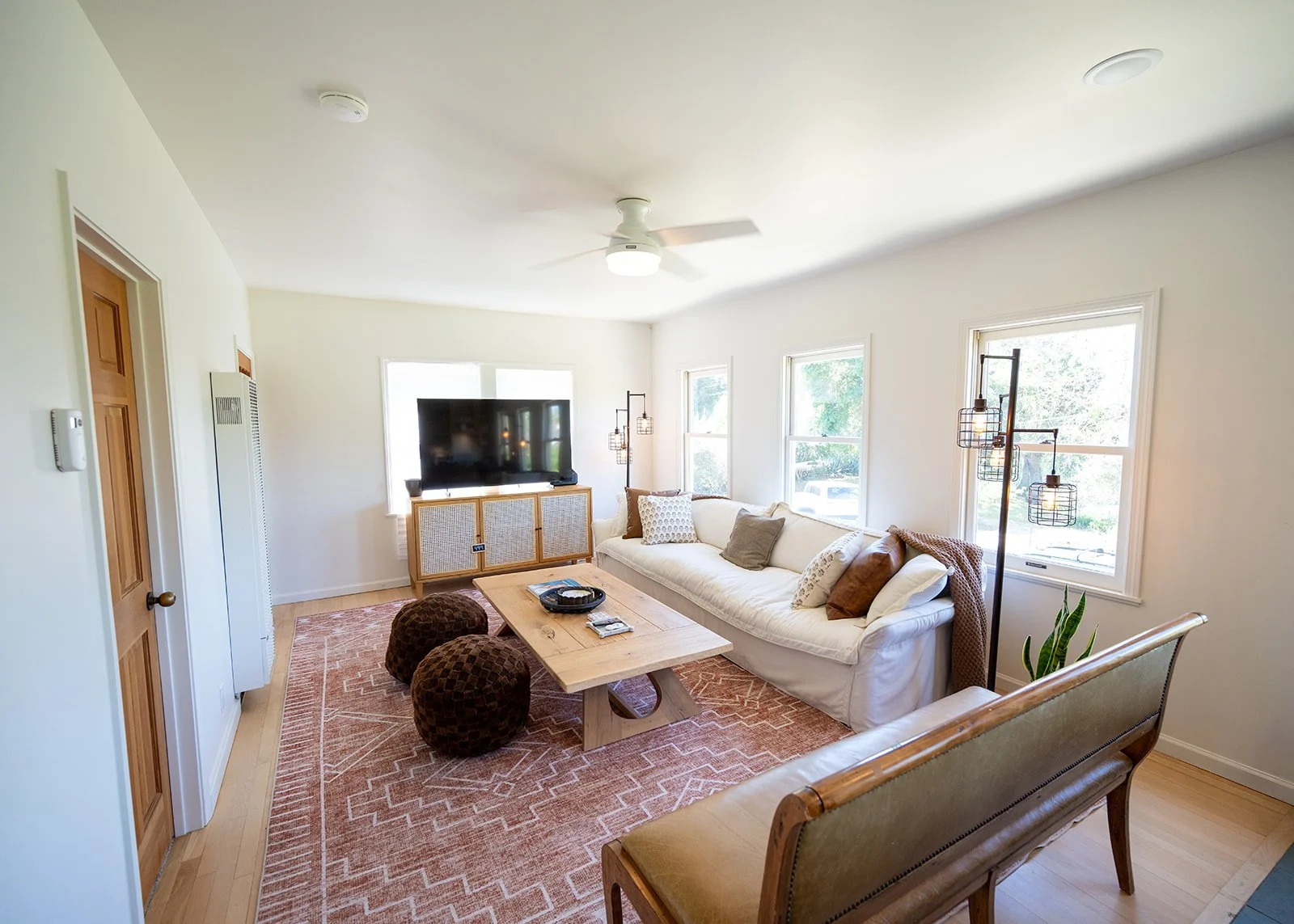 Bright living room with a white sofa, wooden coffee table, large flat-screen TV, multiple windows, and various lamps on a patterned rug.