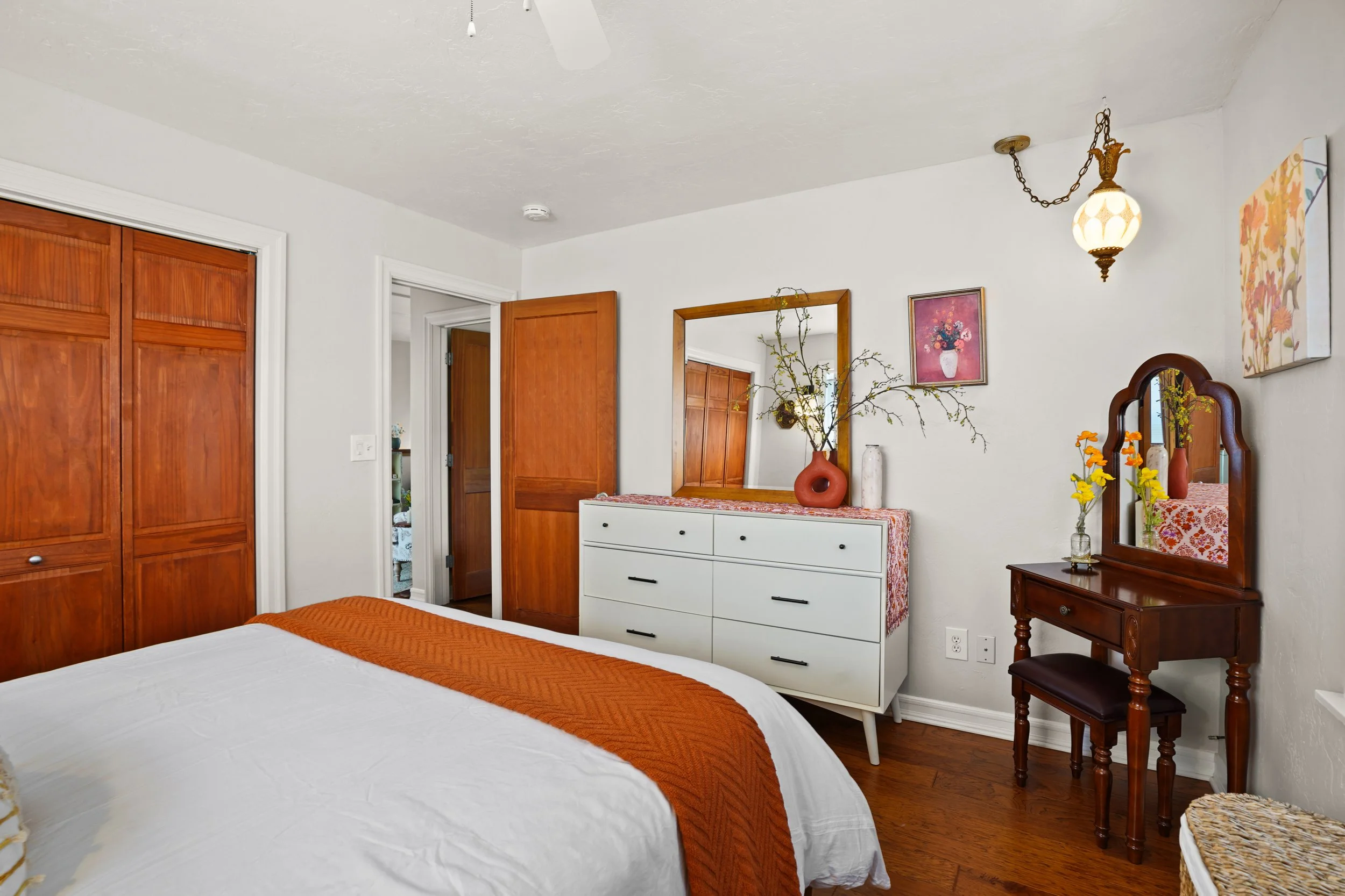 A bedroom with a bed, dresser, mirror, and wooden wardrobe. The bed has white bedding and an orange throw. Decorations include vases with flowers, framed artwork, and a vintage light fixture.