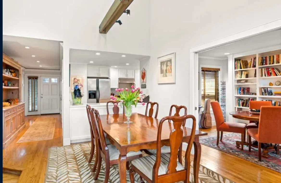 A dining room with a wooden table and chairs, decorated with a vase of pink tulips, adjacent to a kitchen with white cabinets, and a living space with orange chairs and bookshelves.