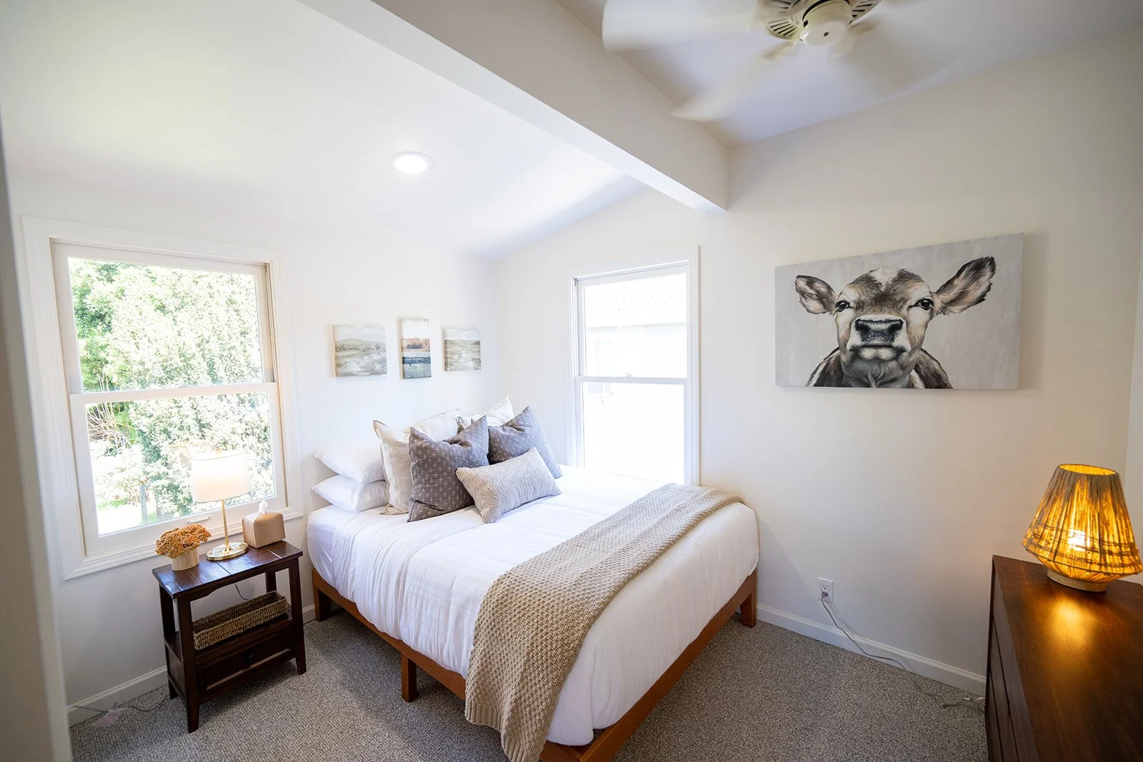 Bright bedroom with white walls, a bed with multiple pillows, a nightstand with a lamp and decorative flowers, wall art including a cow painting and three landscape pictures, two windows, and a ceiling fan.