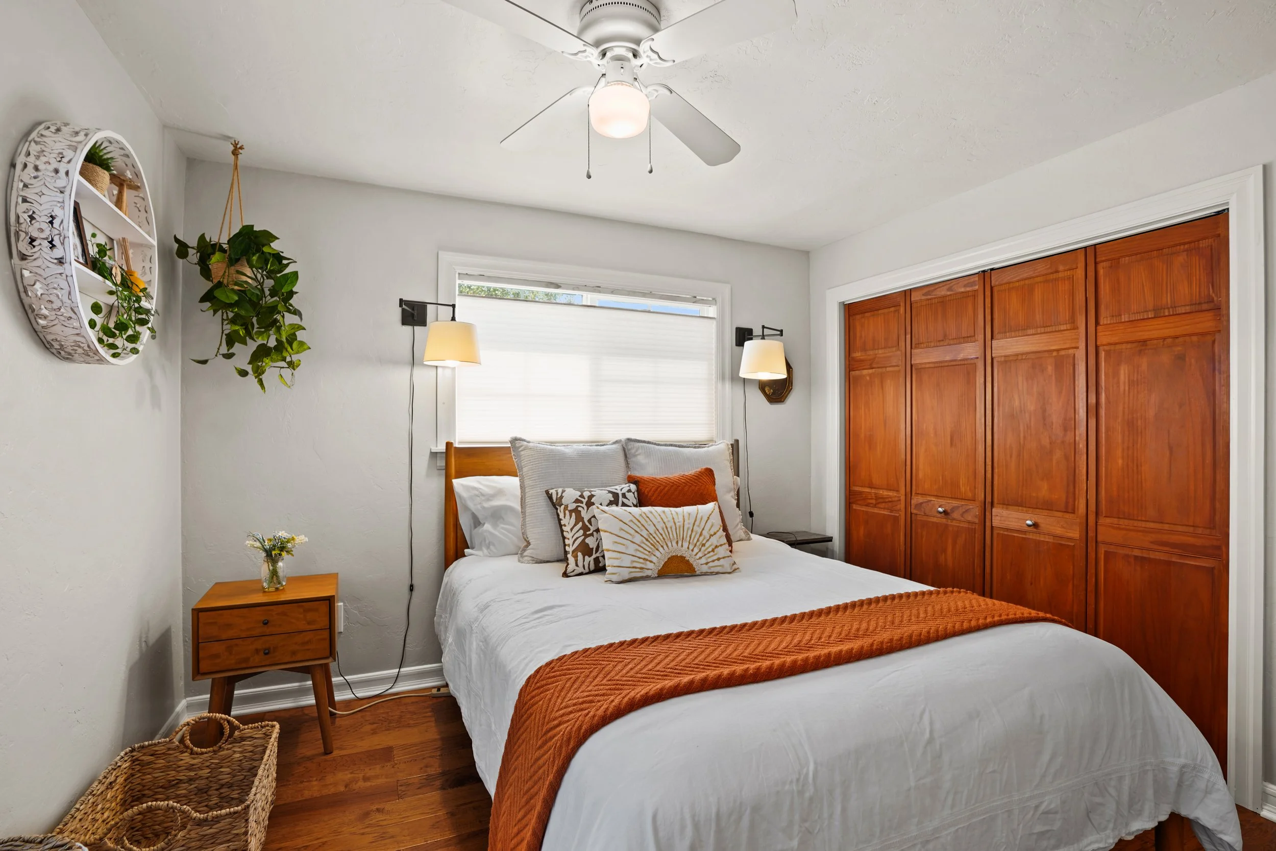 A cozy bedroom with a white bed, patterned cushions, and an orange blanket. Nightstands on each side with lamps, a window with white blinds, a wooden closet door, wall-mounted lights, a ceiling fan, and decorative wall shelves with plants and photos.