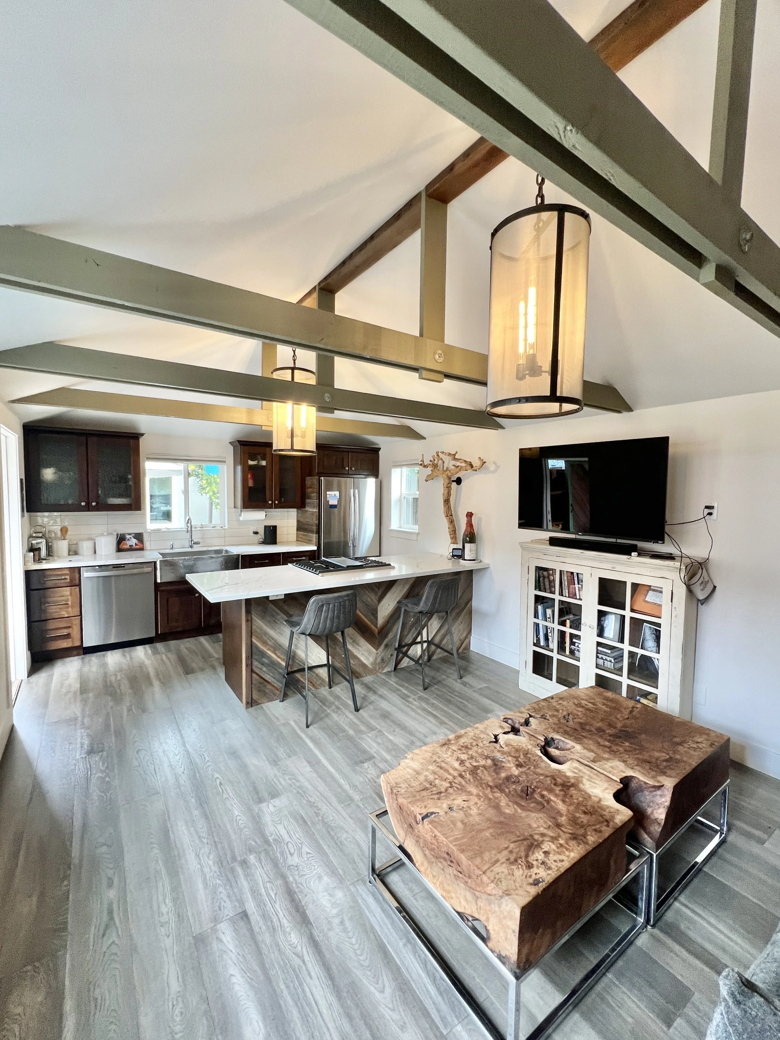 Open-concept kitchen and living room with vaulted ceiling, modern lighting, dark wood cabinetry, stainless steel appliances, a white marble kitchen island with two bar stools, a TV on a white cabinet, and a rustic wooden coffee table.