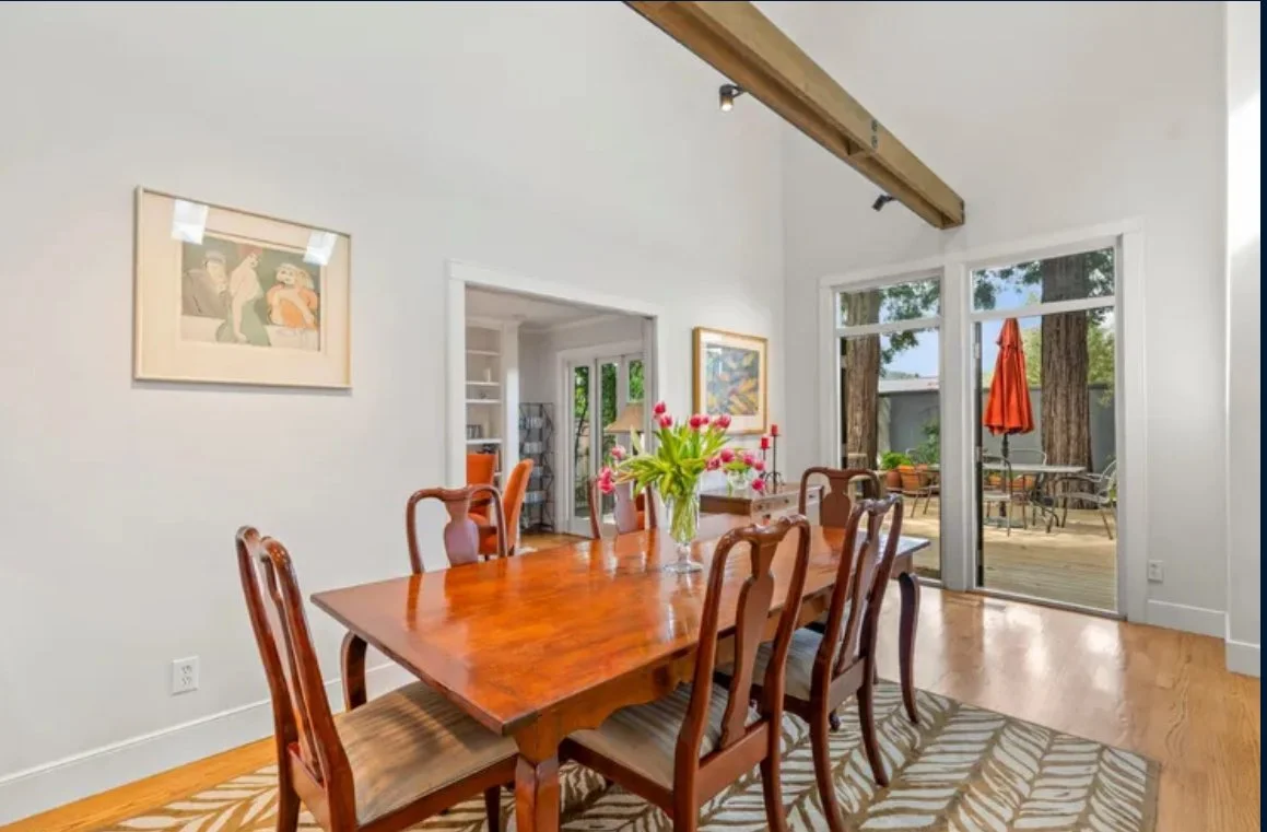 Dining room with a wooden table and chairs, a vase of pink tulips, and a view of an outdoor patio with a table, chairs, and an umbrella.
