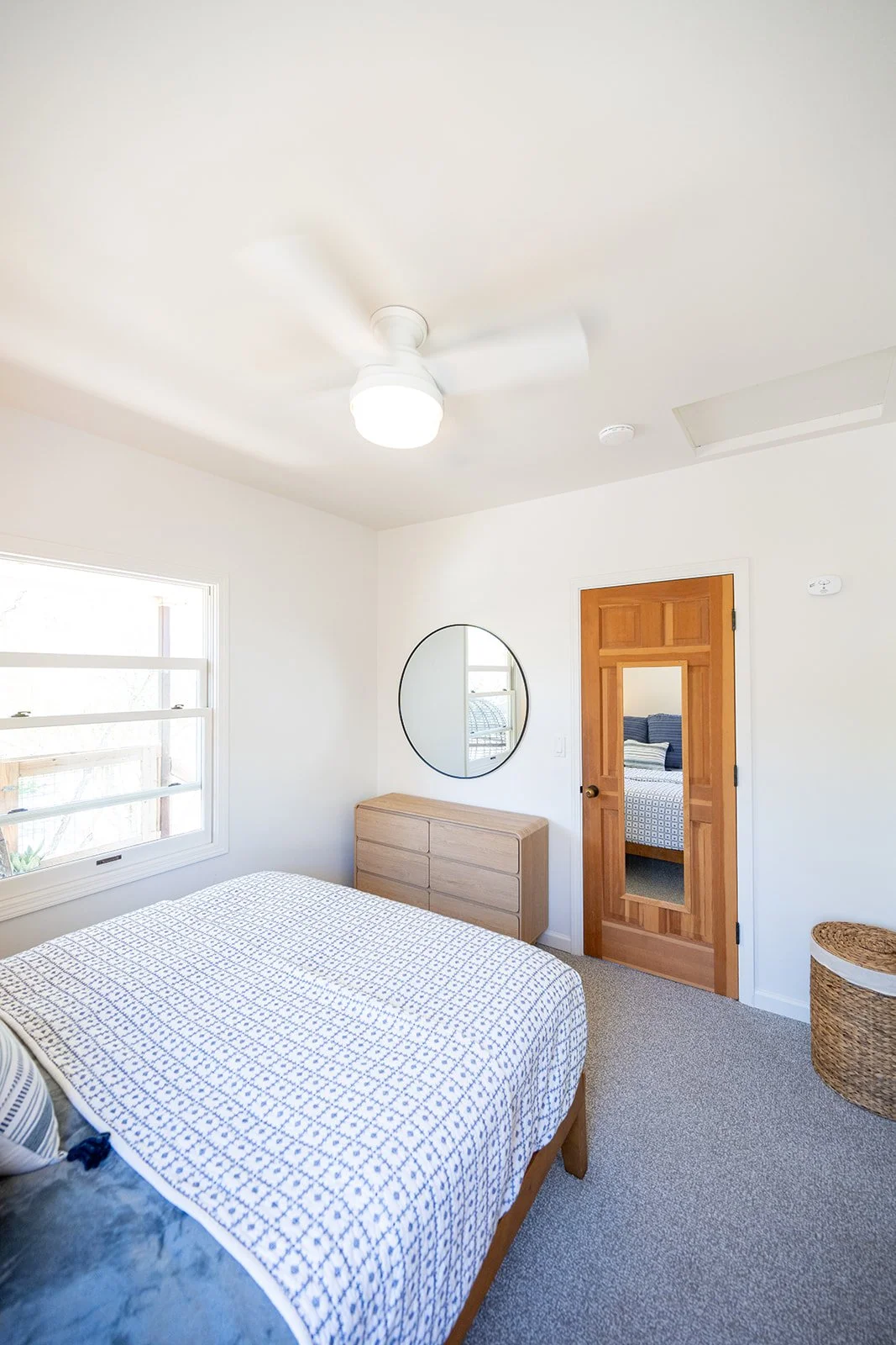A minimalist bedroom with a bed, a wooden dresser, a round mirror on the wall, a window, a wooden door with a mirror, and a wicker laundry basket in a bright room.