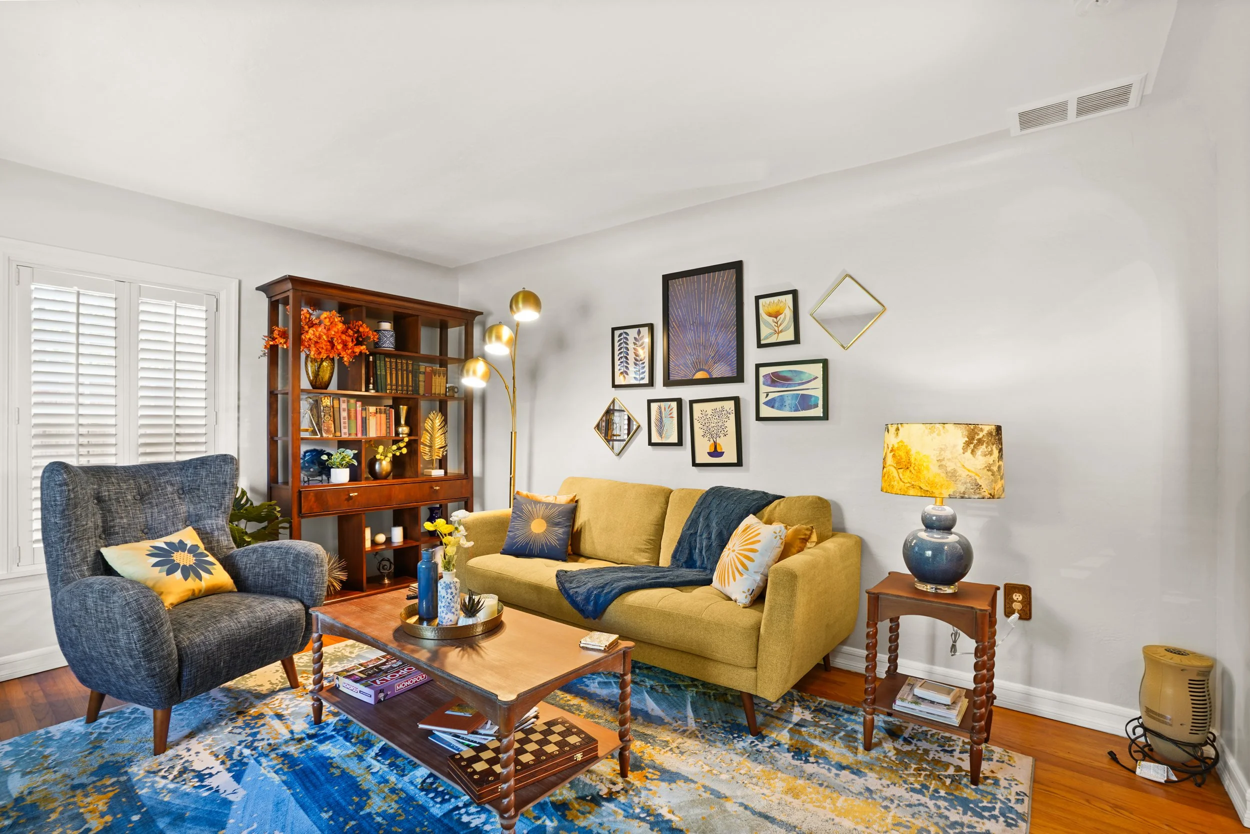 Living room with a yellow sofa, a gray armchair, and decorative pillows. Wall art and a bookshelf with plants and books. A wooden coffee table with a tray of decor items, and a blue patterned rug. A lamp with a floral shade on a side table.