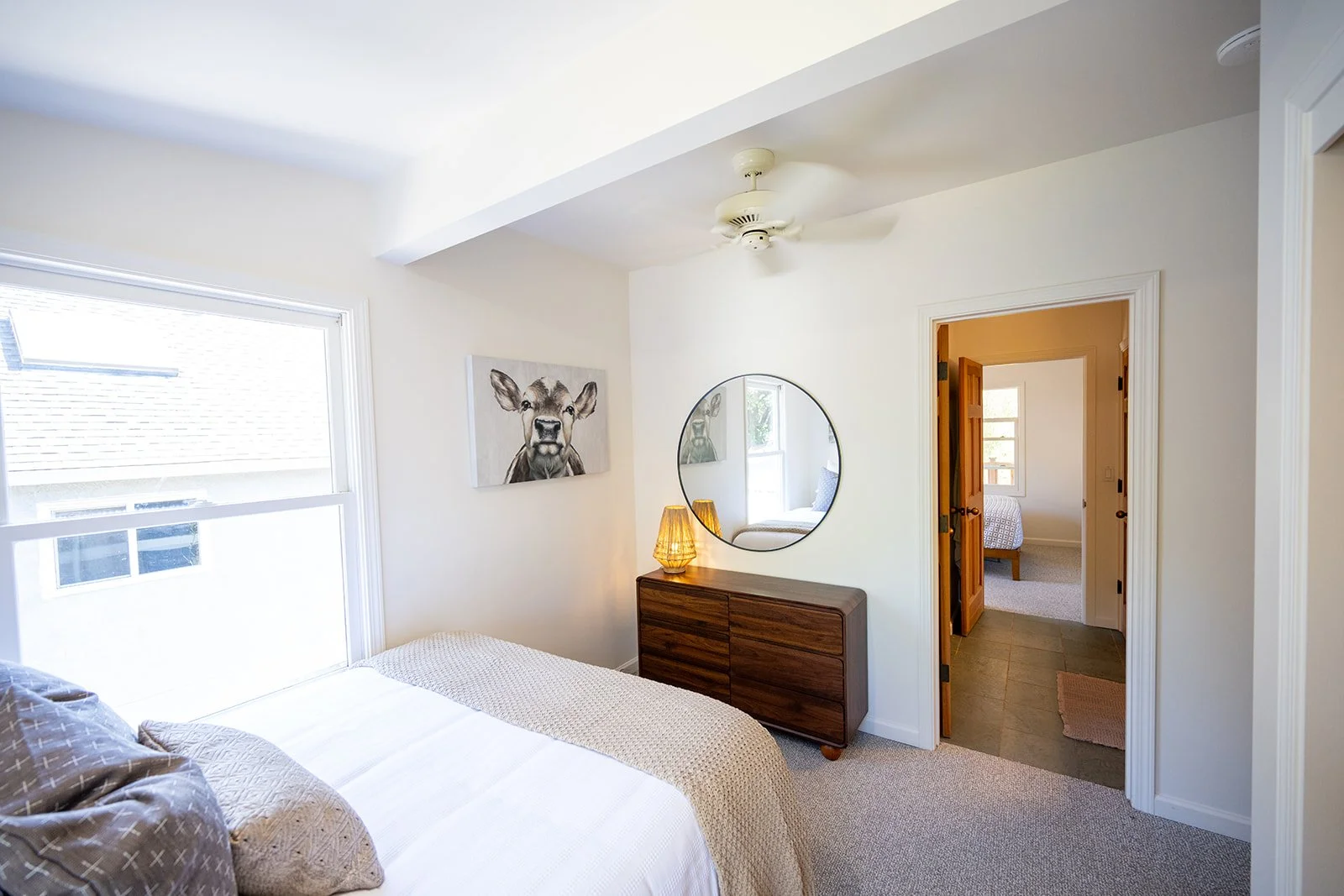 A cozy bedroom with a large window, a bed with beige and gray pillows, a wall art of a cow, a round mirror, a wooden dresser with two lamps, and a doorway leading to another room with a bed, tiled and carpeted flooring, and a window.