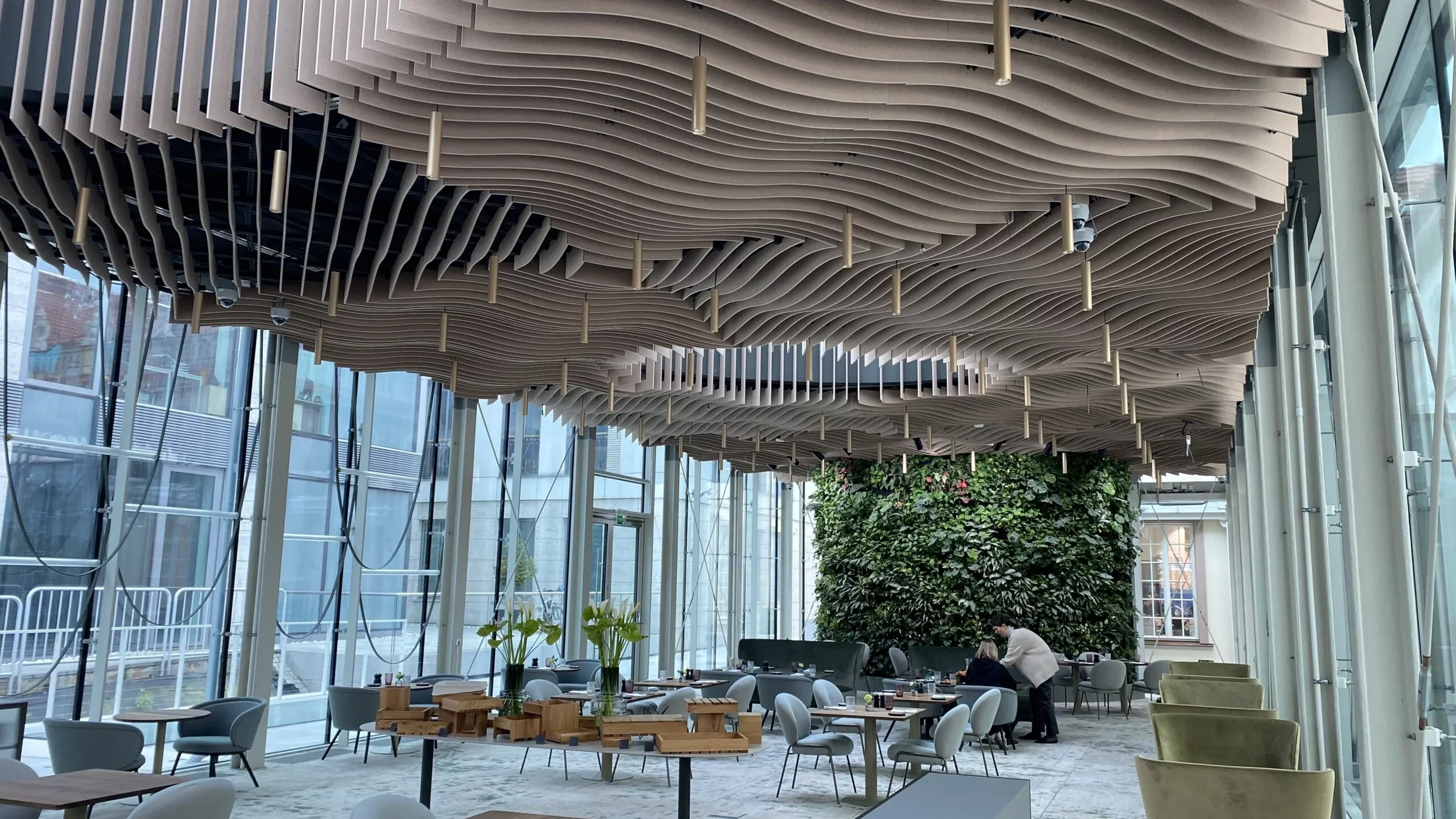 A modern indoor space with a wave-patterned wooden ceiling, large glass windows, and a living green wall. There are tables, chairs, and people seated and standing, suggesting a restaurant or cafe setting.