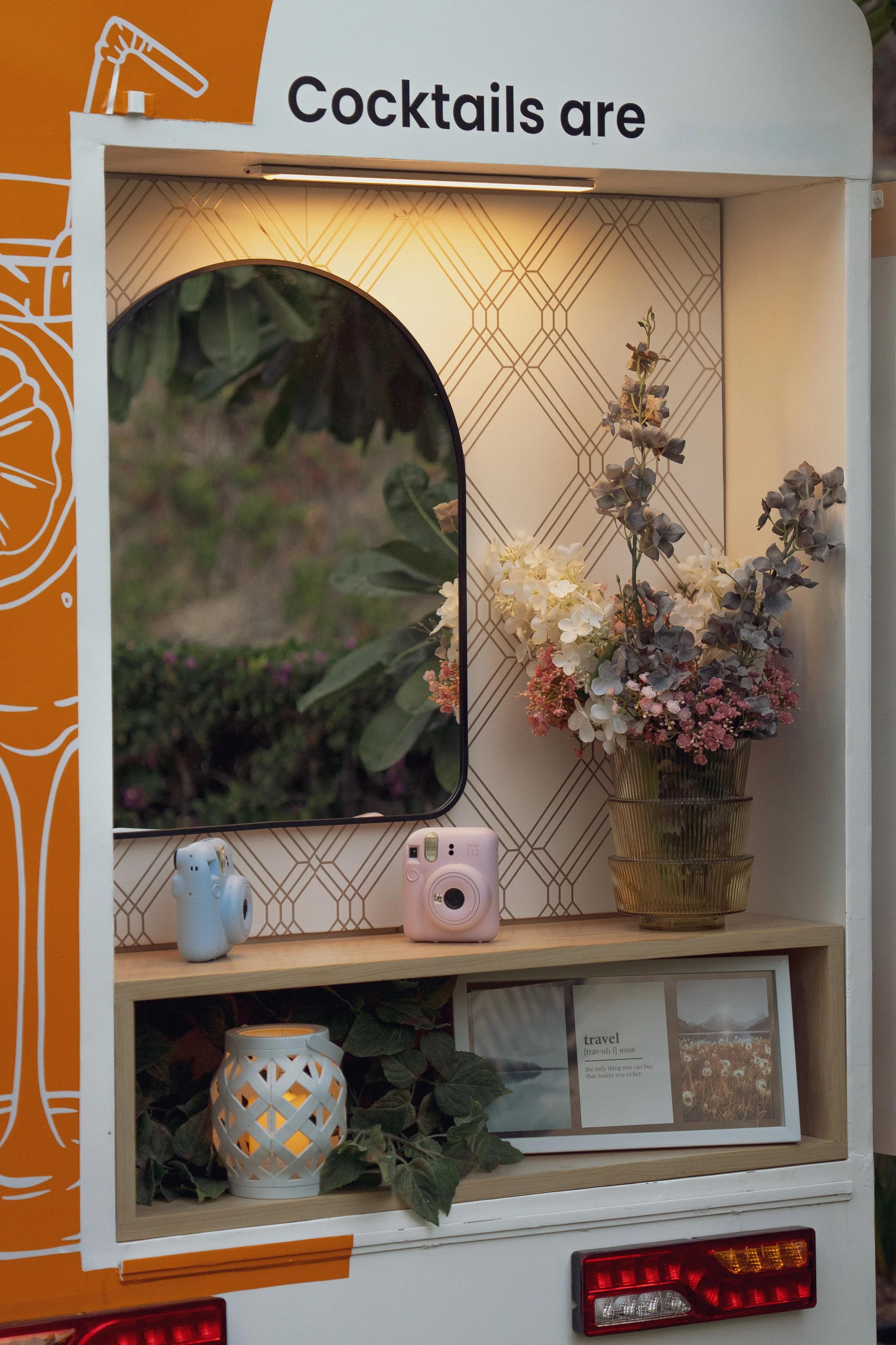 A colorful photo of a mobile cocktail cart with a sign reading 'Cocktails are'. The cart has a round mirror, a pink instant camera, and a blue camera on the shelf. There is a vase with mixed flowers and a decorative white candle holder with green plants, and photo prints below.