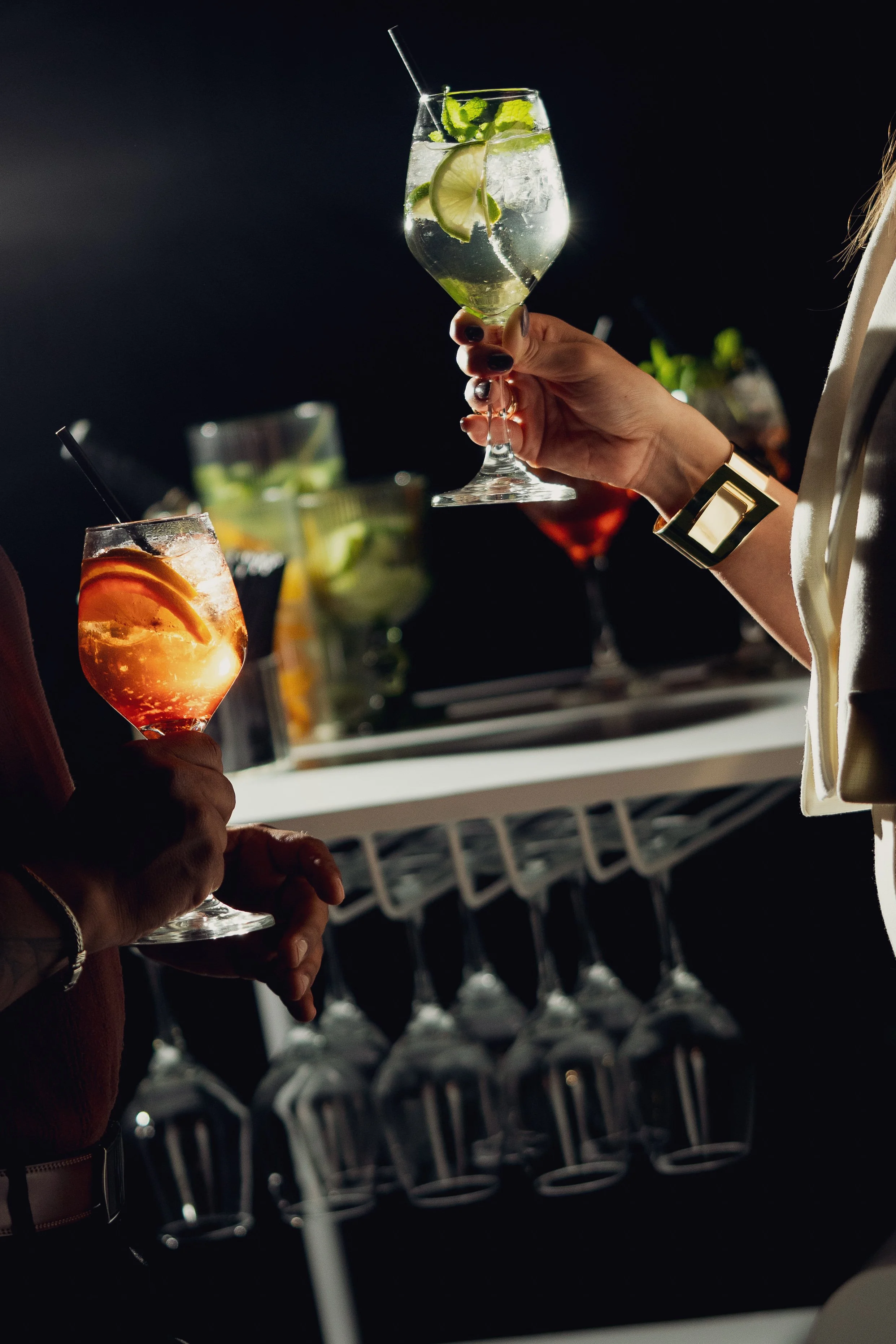 Two people hold cocktails, one with a lemon slice and mint leaves, in a dark setting, possibly a bar, with glasses and bottles in the background.