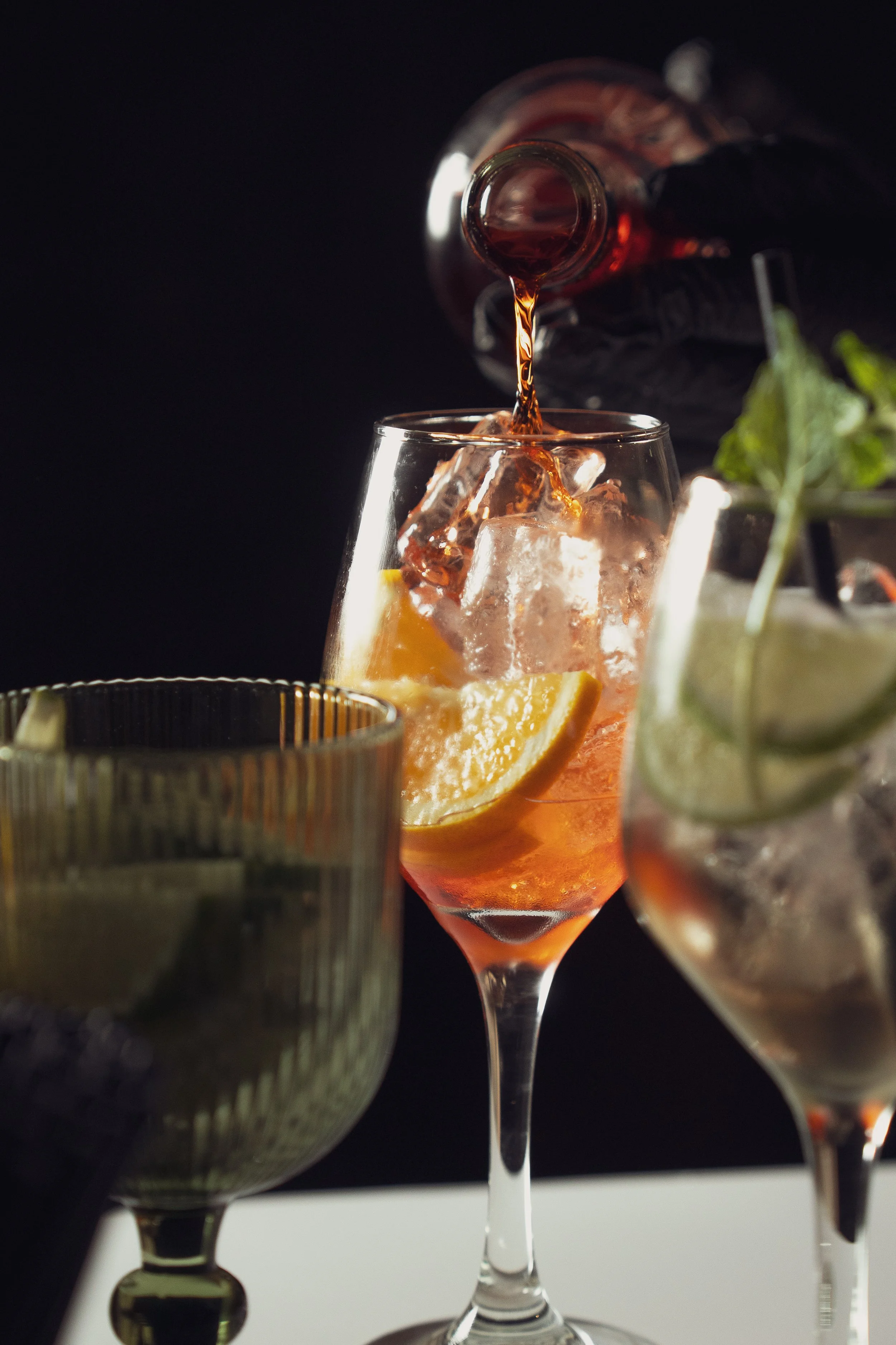 A cocktail glass being filled with a dark beverage from a bottle, with ice cubes and lemon slices inside the glass, against a dark background.