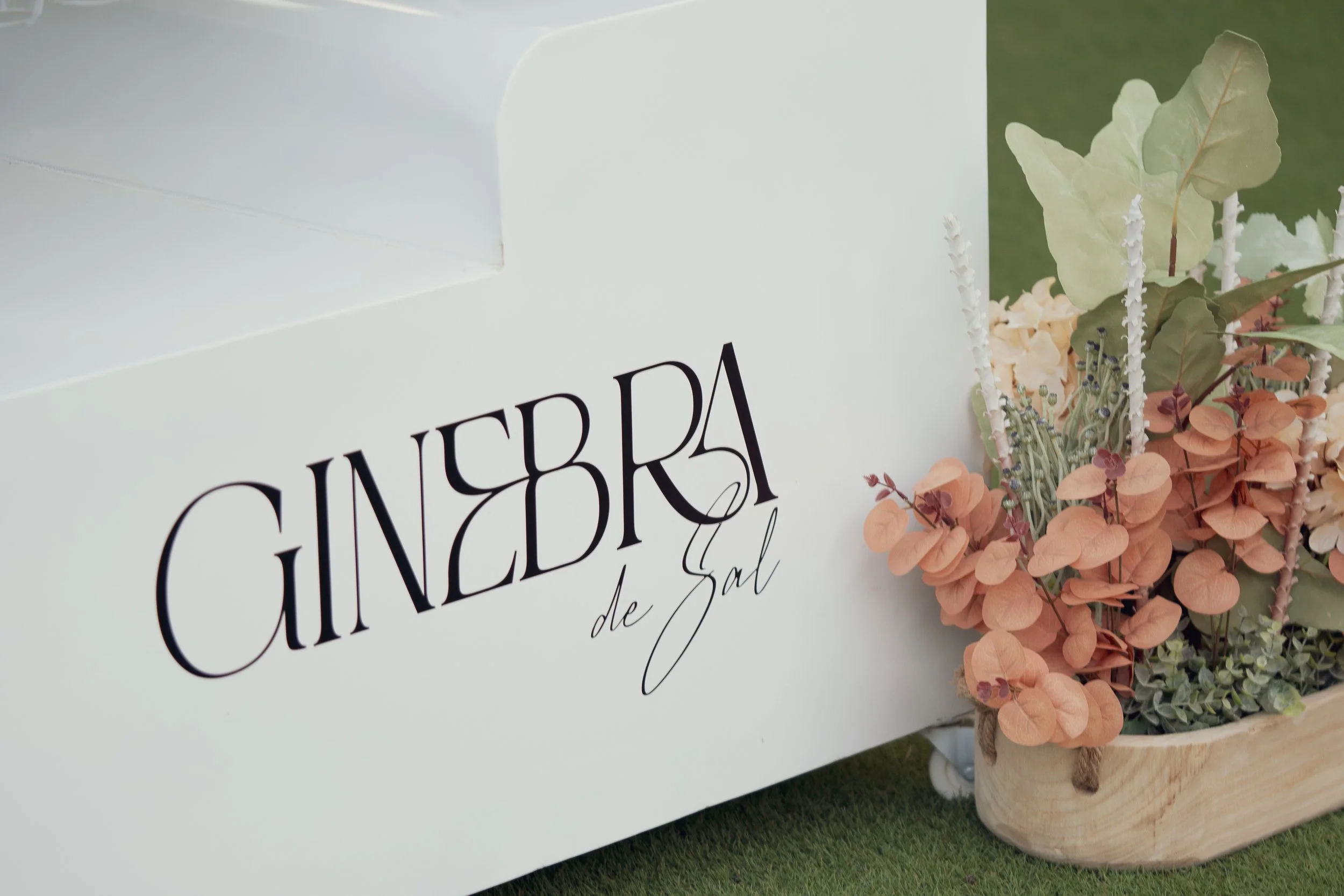 White flower cart with black lettering and a floral arrangement of peach, green, and purple flowers and greenery