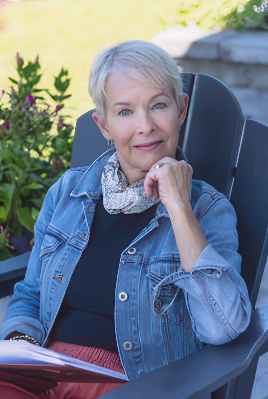 A smiling woman with short blonde hair, wearing a red blazer over a black and white animal print turtleneck, against a black background.