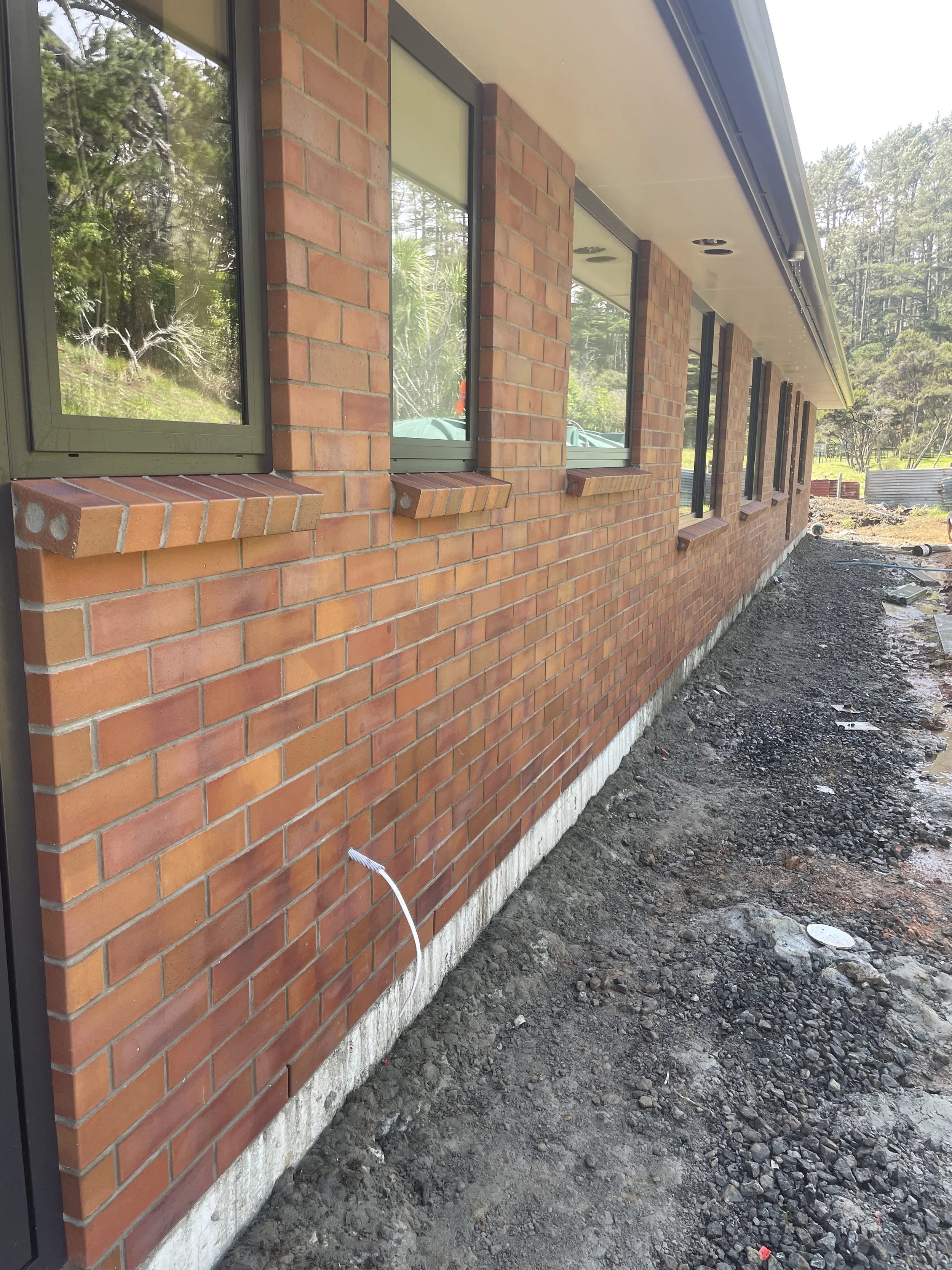 Exterior view of a building under construction with a brick wall and multiple windows, construction materials on the ground, and a forested area in the background.
