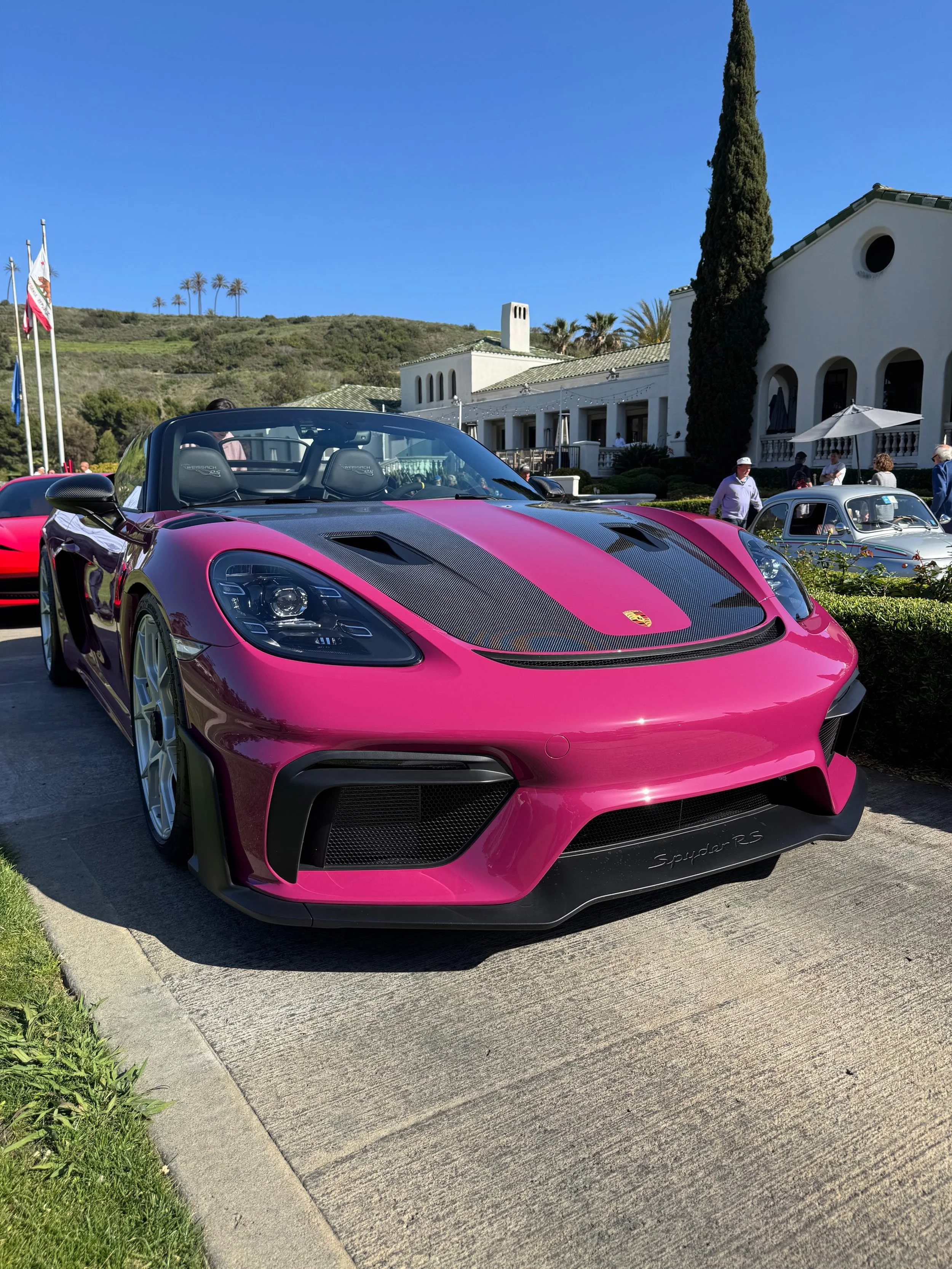 Ruby Star Porsche Spyder RS parked outdoors with a white building, palm trees, and a cypress tree in the background on a clear, sunny day.