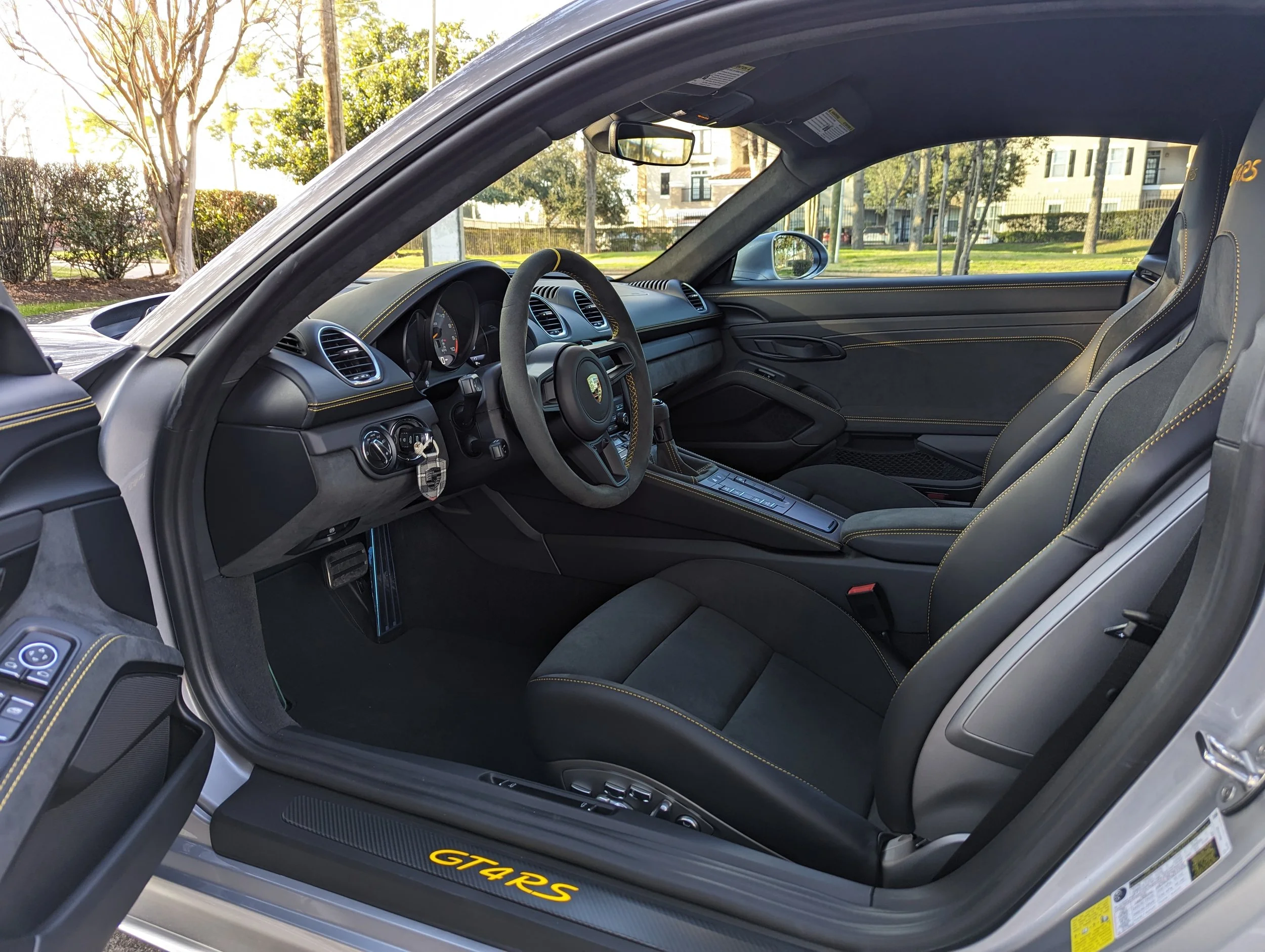 Interior of a Porsche 911 GT4 RS showing black leather seats with yellow stitching, steering wheel, dashboard, and center console, with a view of trees and buildings outside