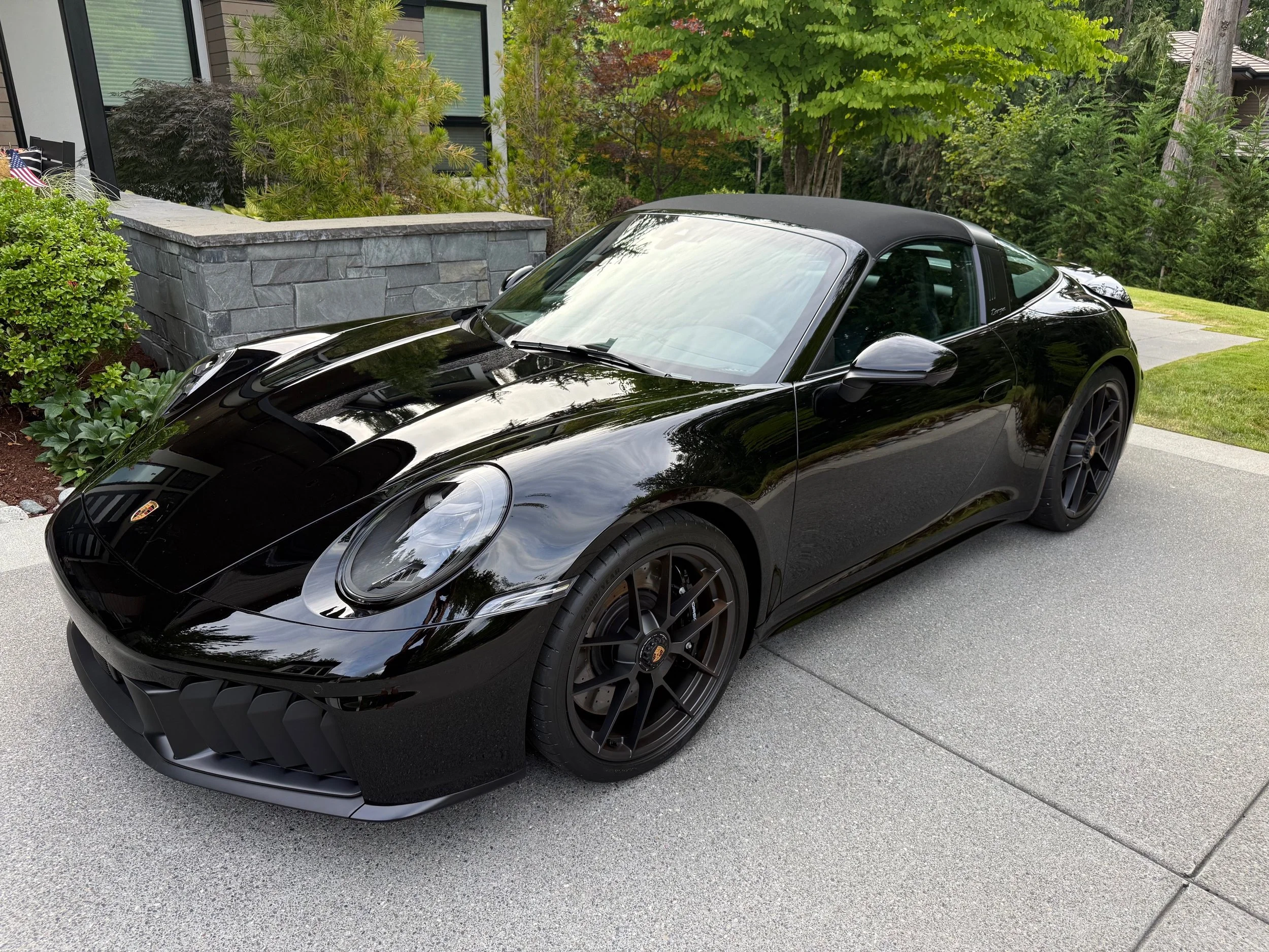 Black luxury convertible sports car parked on driveway next to house with flowers and trees in the background.