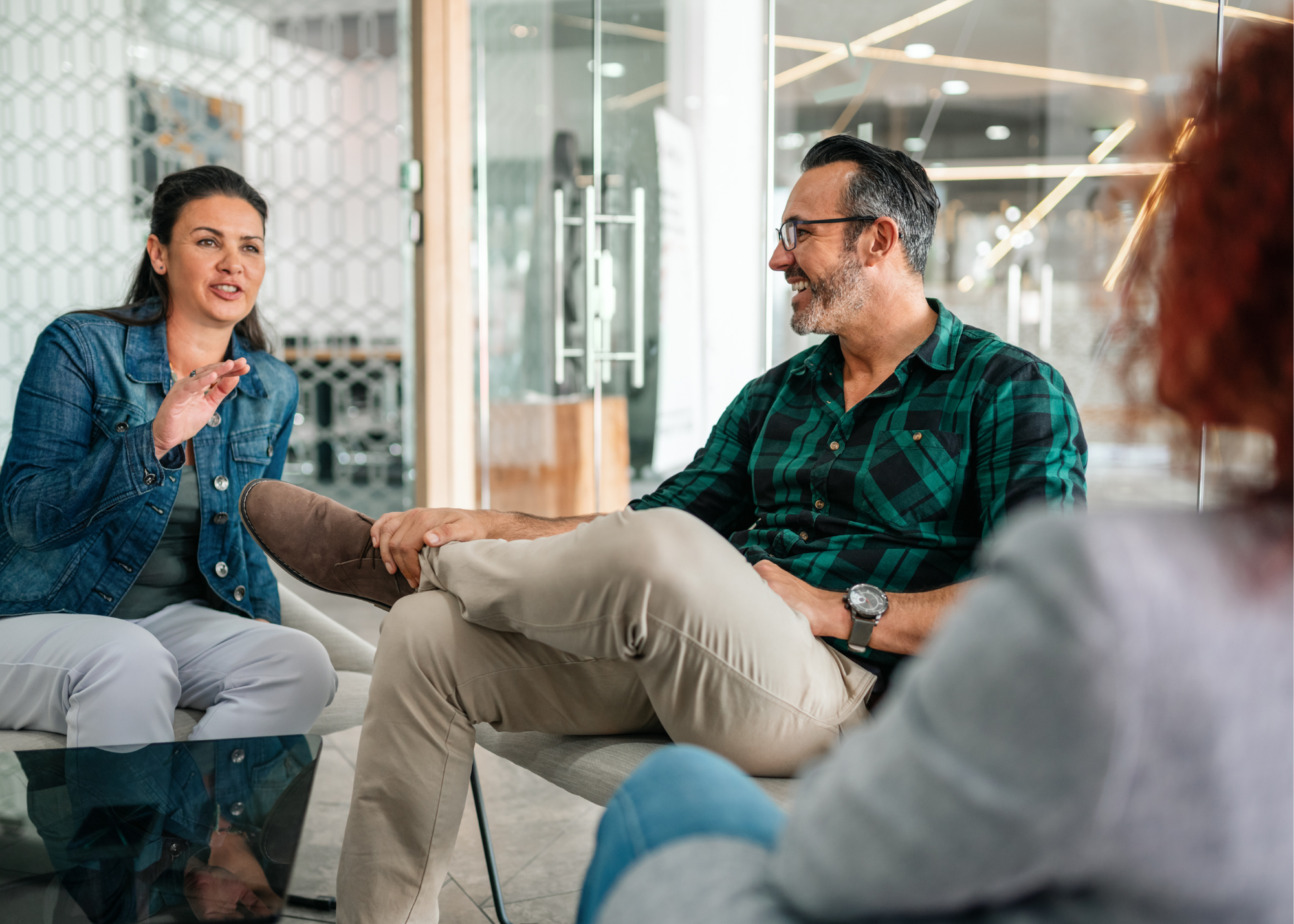 Three people having a conversation in a modern indoor setting. One woman is speaking, while a man is smiling and listening, with another person partly visible in the foreground.