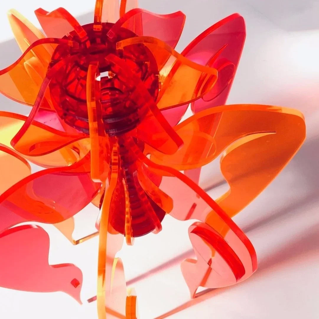 A colorful, abstract sculpture resembling a flower, made of translucent orange and red plastic pieces assembled with a black base.